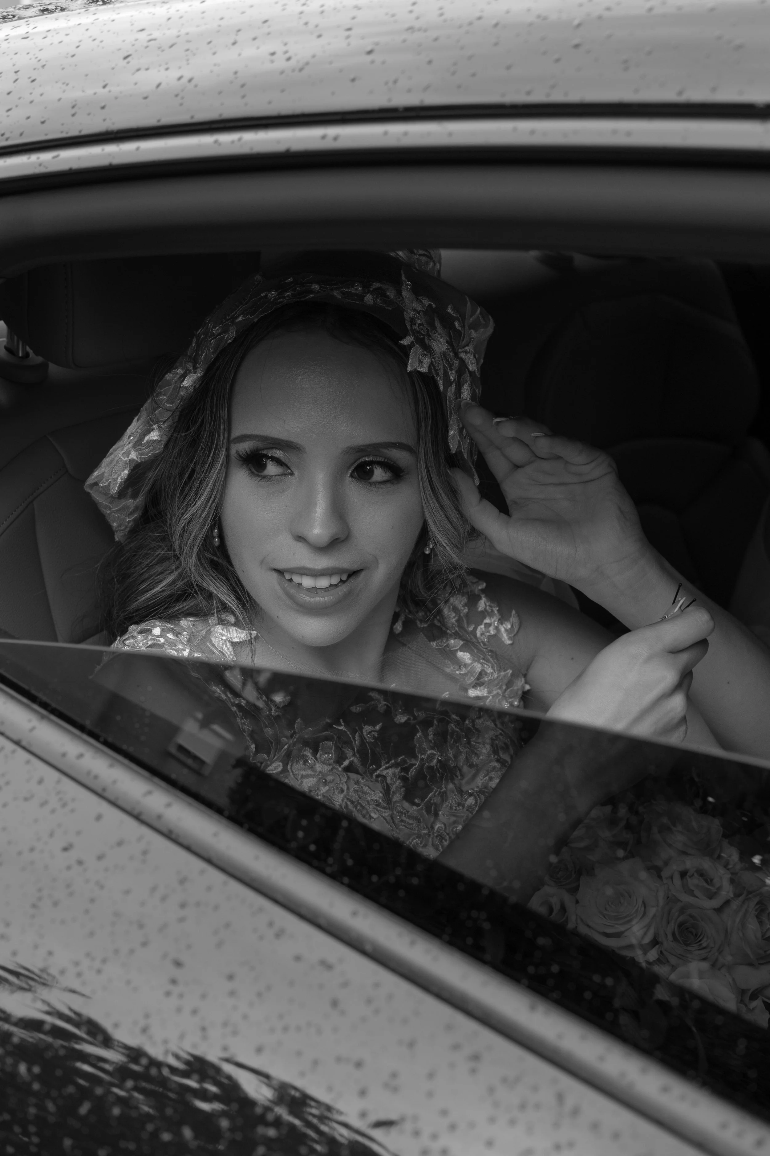 A woman wearing a lace headscarf and earrings sitting in a car, looking out the window with a slight smile, with a bouquet of roses visible inside the vehicle.