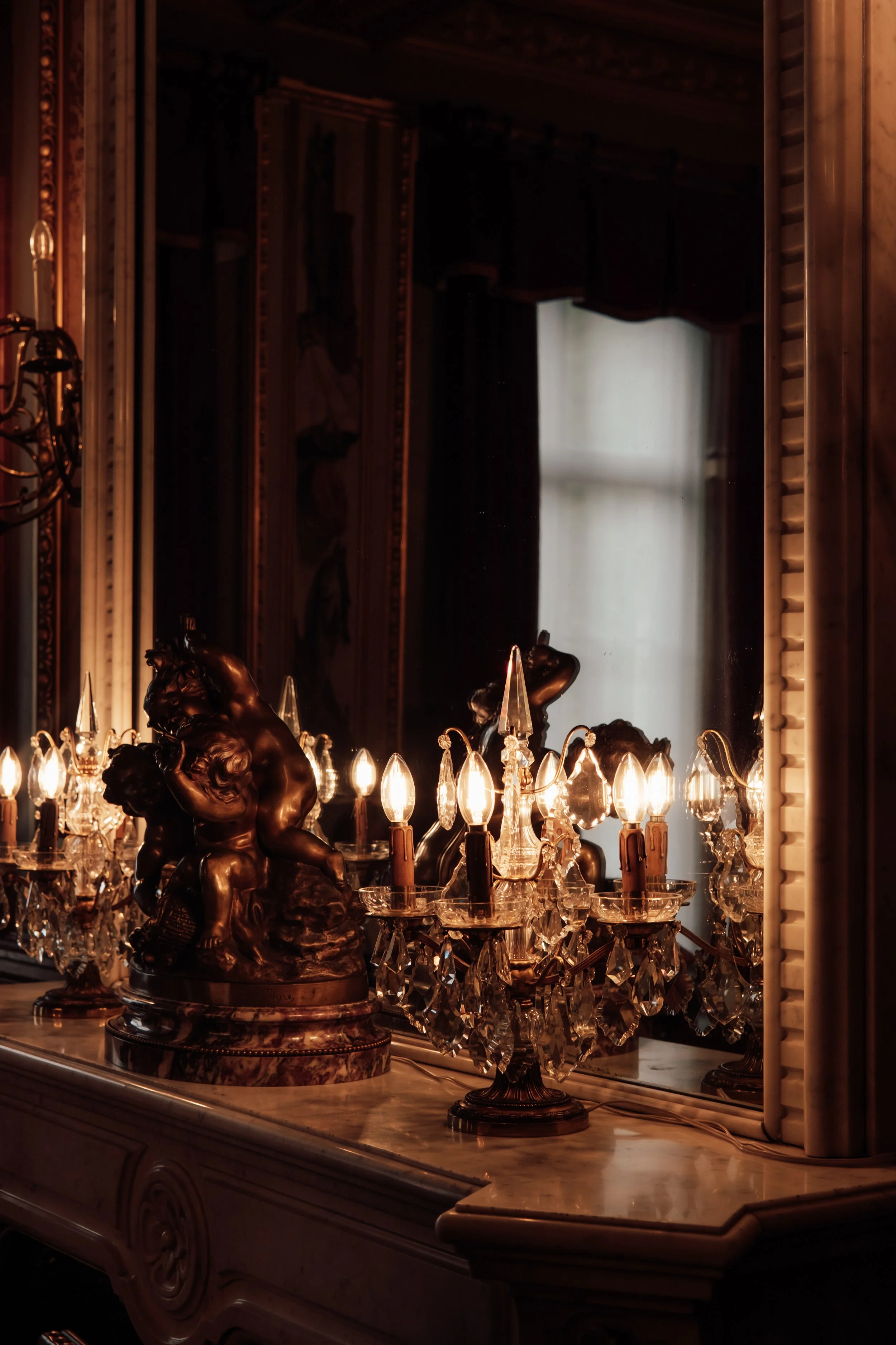 Vintage chandelier with crystal accents and a bronze cherub sculpture on the ornate wooden table in a dimly lit room with dark curtains and a window in the background.