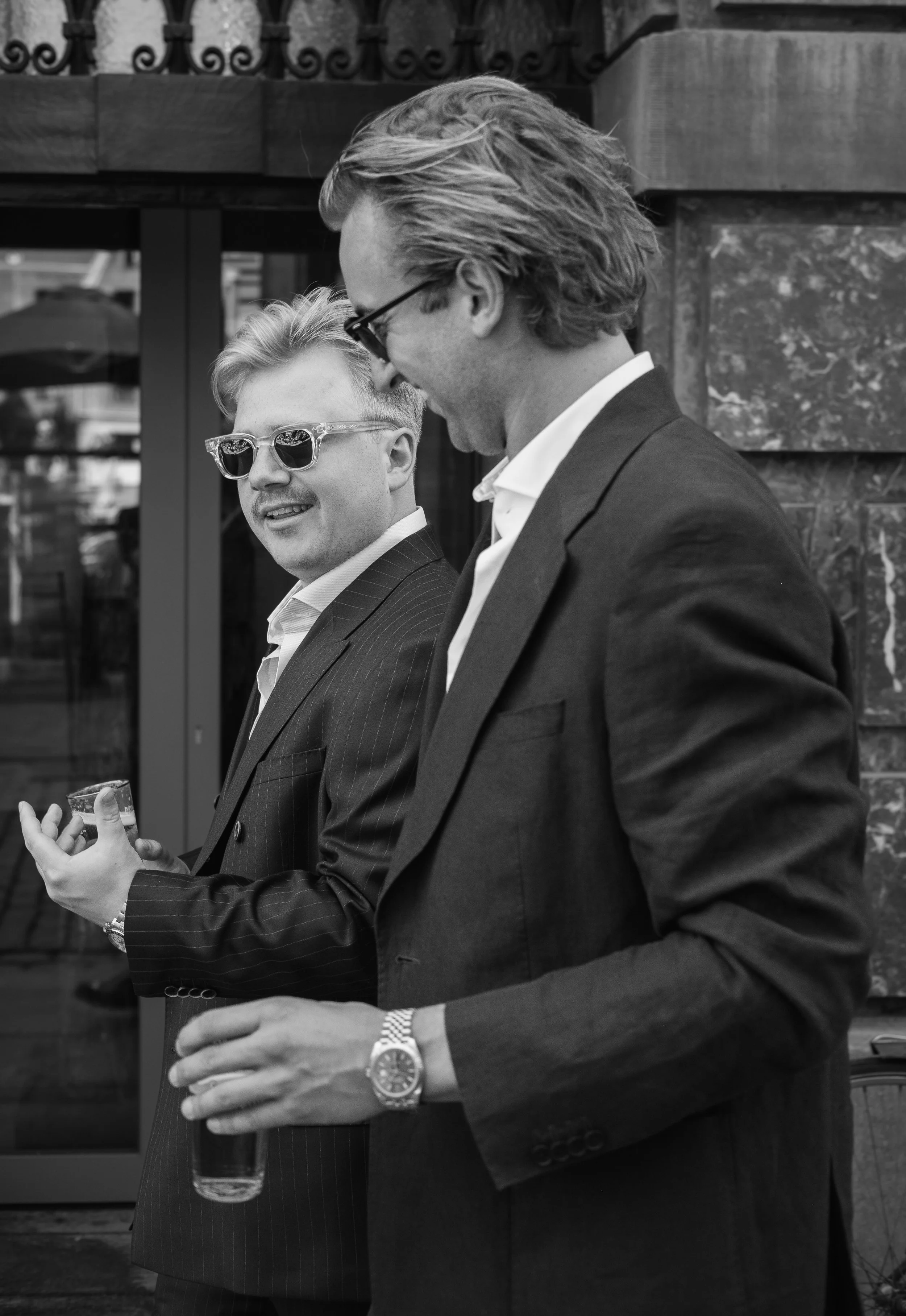 Two men in suits talking to each other, one wearing sunglasses and the other with glasses, at an outdoor event, with one holding a glass of drink.