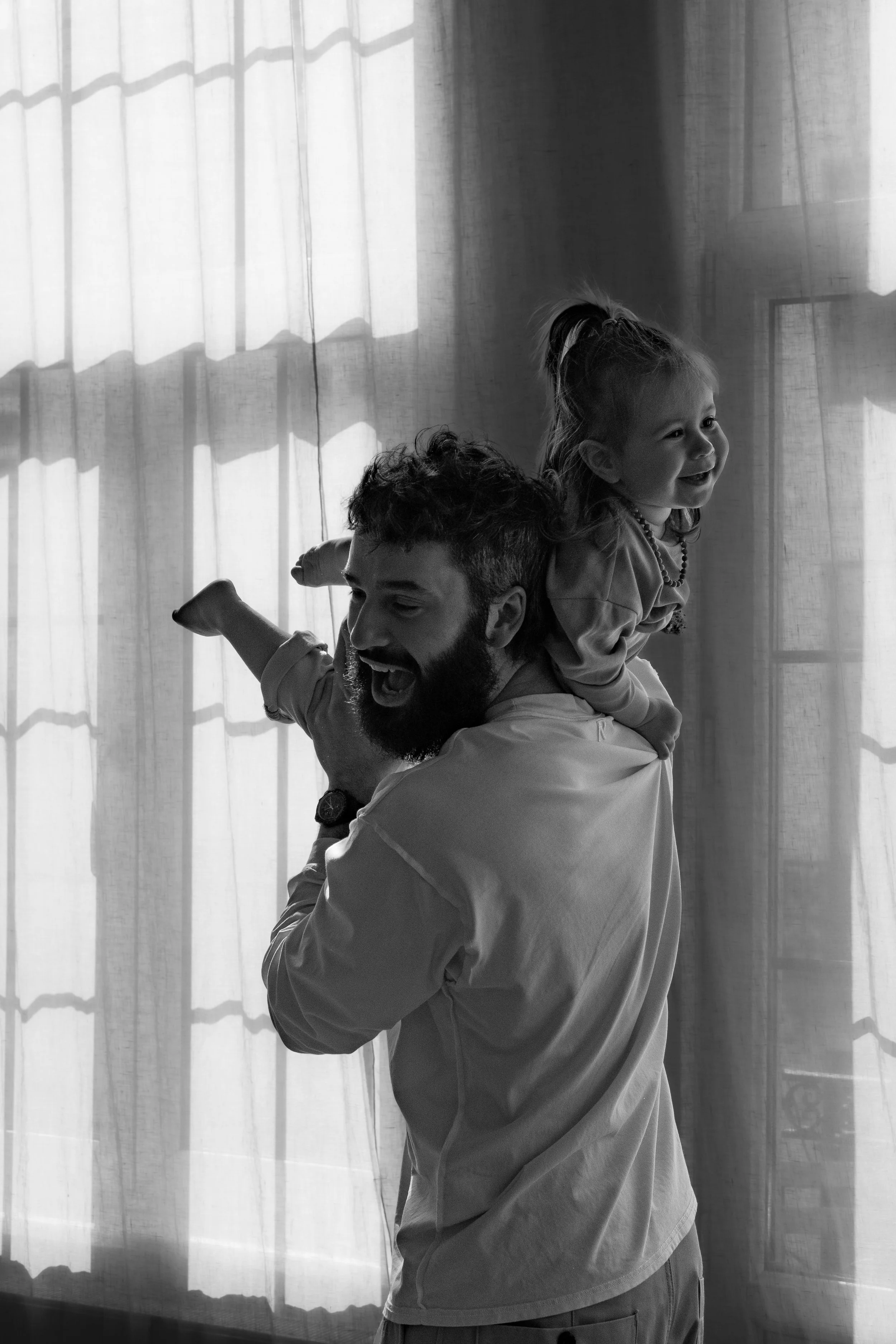A man with a beard is carrying a young girl on his shoulders indoors in front of sheer curtains. The girl is smiling and looking to the right, while the man is facing left with his eyes closed and mouth open in a joyful expression.