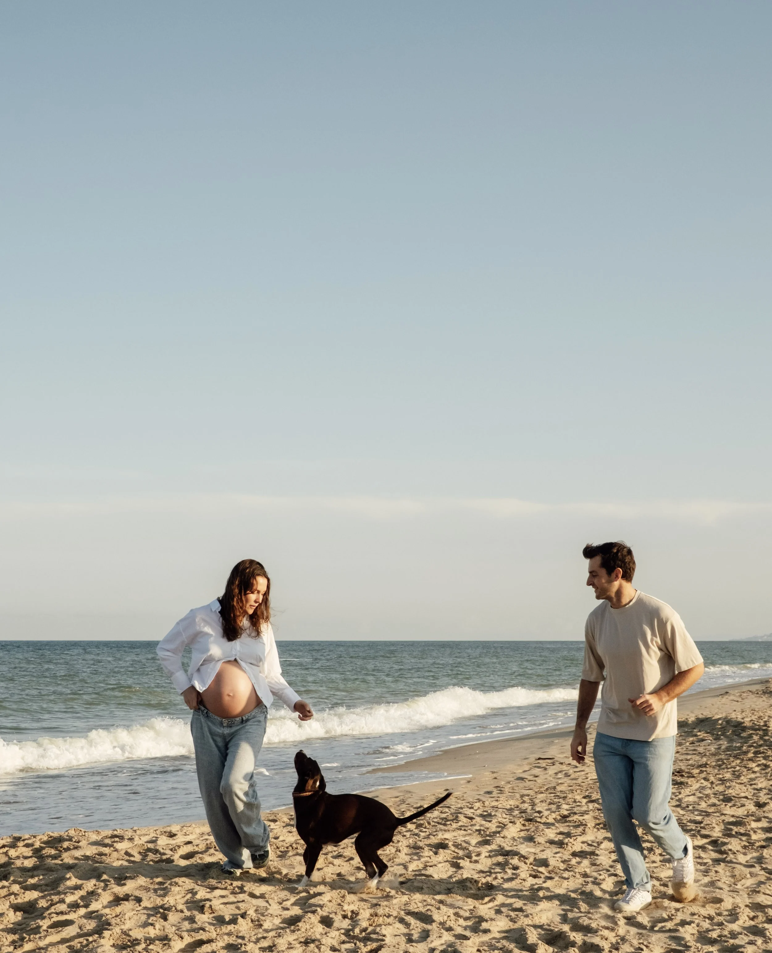 A pregnant woman in a white shirt and jeans walking on the beach with a black dog while a man in a beige shirt and jeans walks beside them.