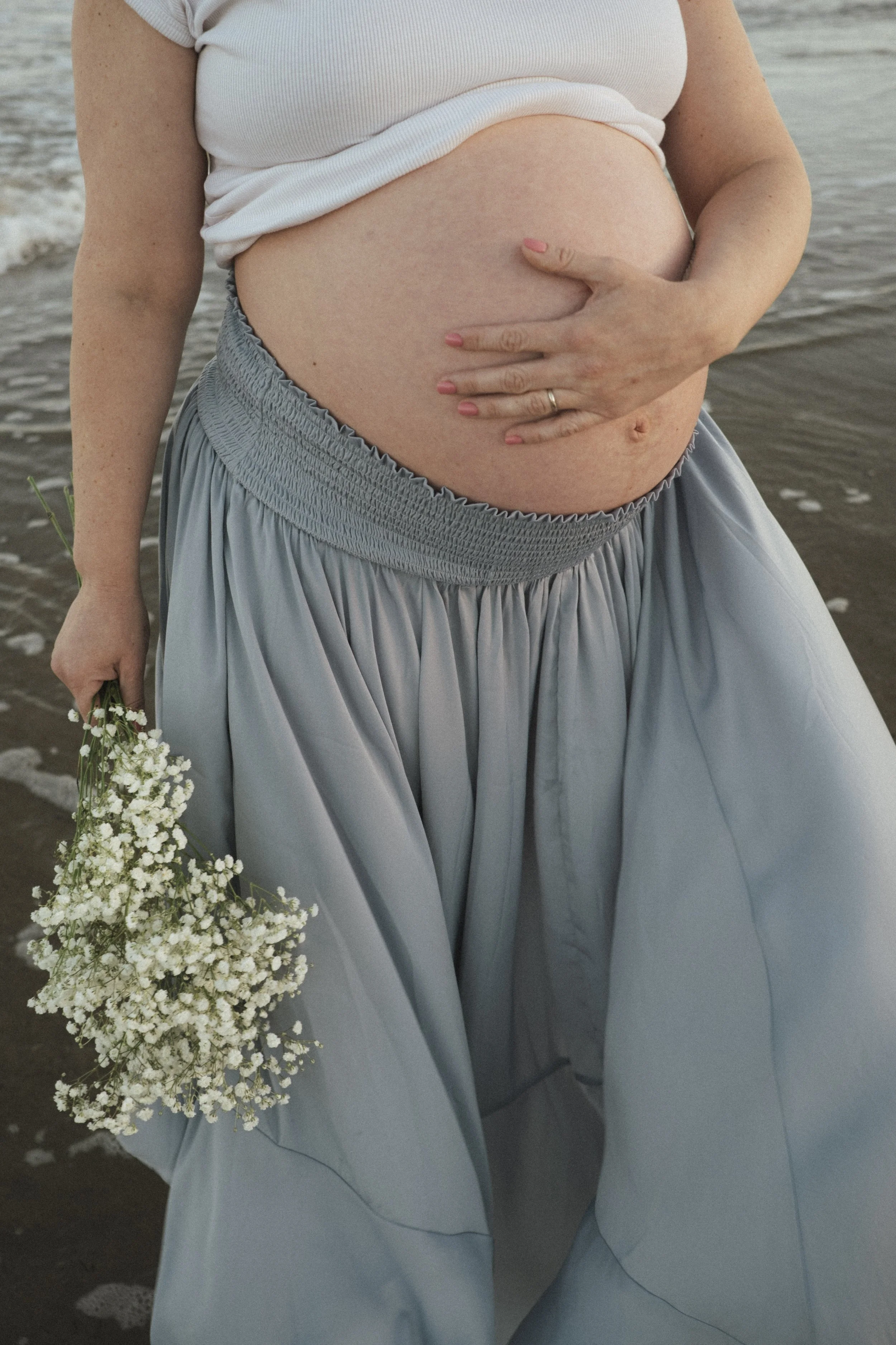 Pregnant person in a white top and blue skirt holding a bouquet of baby's breath flowers.