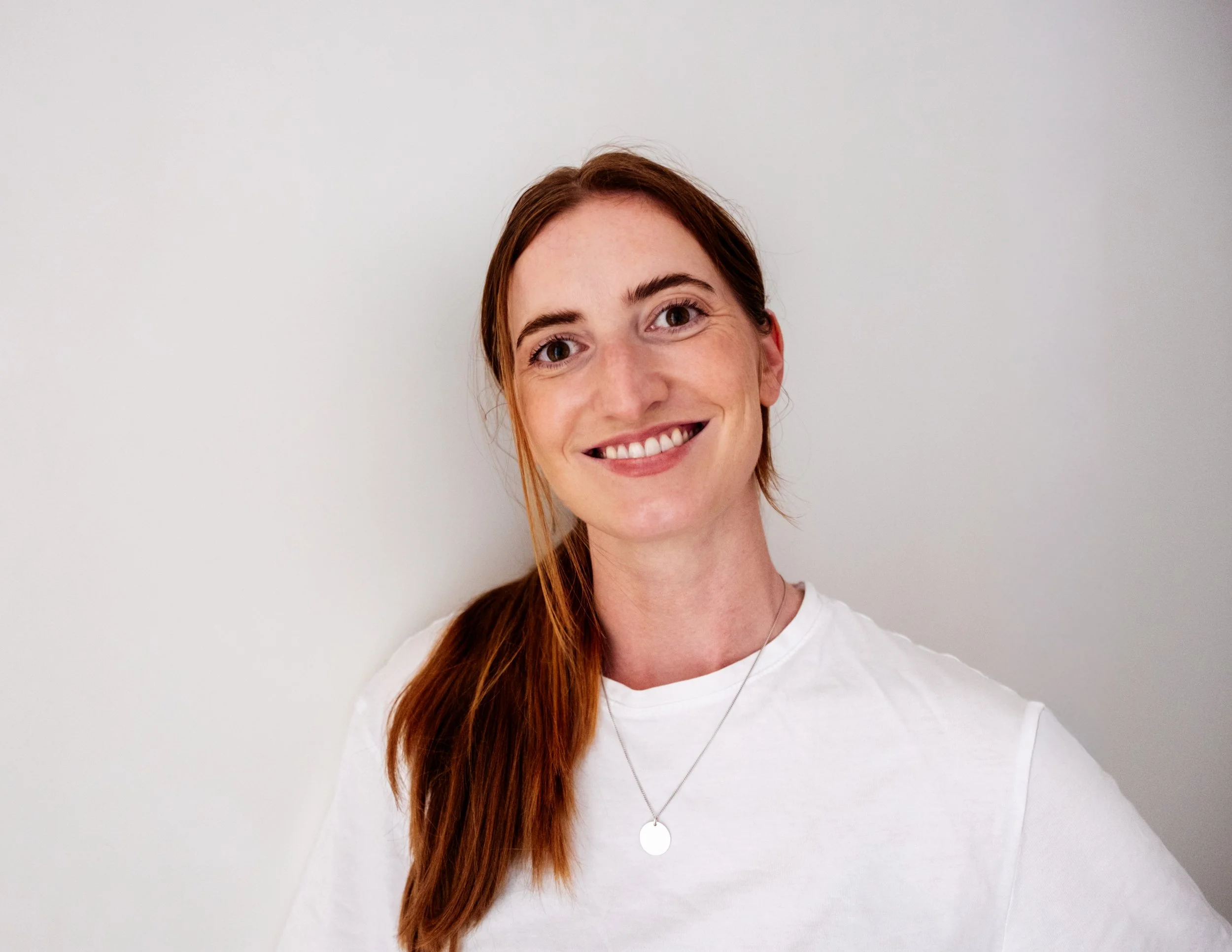 Portrait of a smiling woman with long red hair, wearing a white shirt and a necklace, standing against a plain white background.