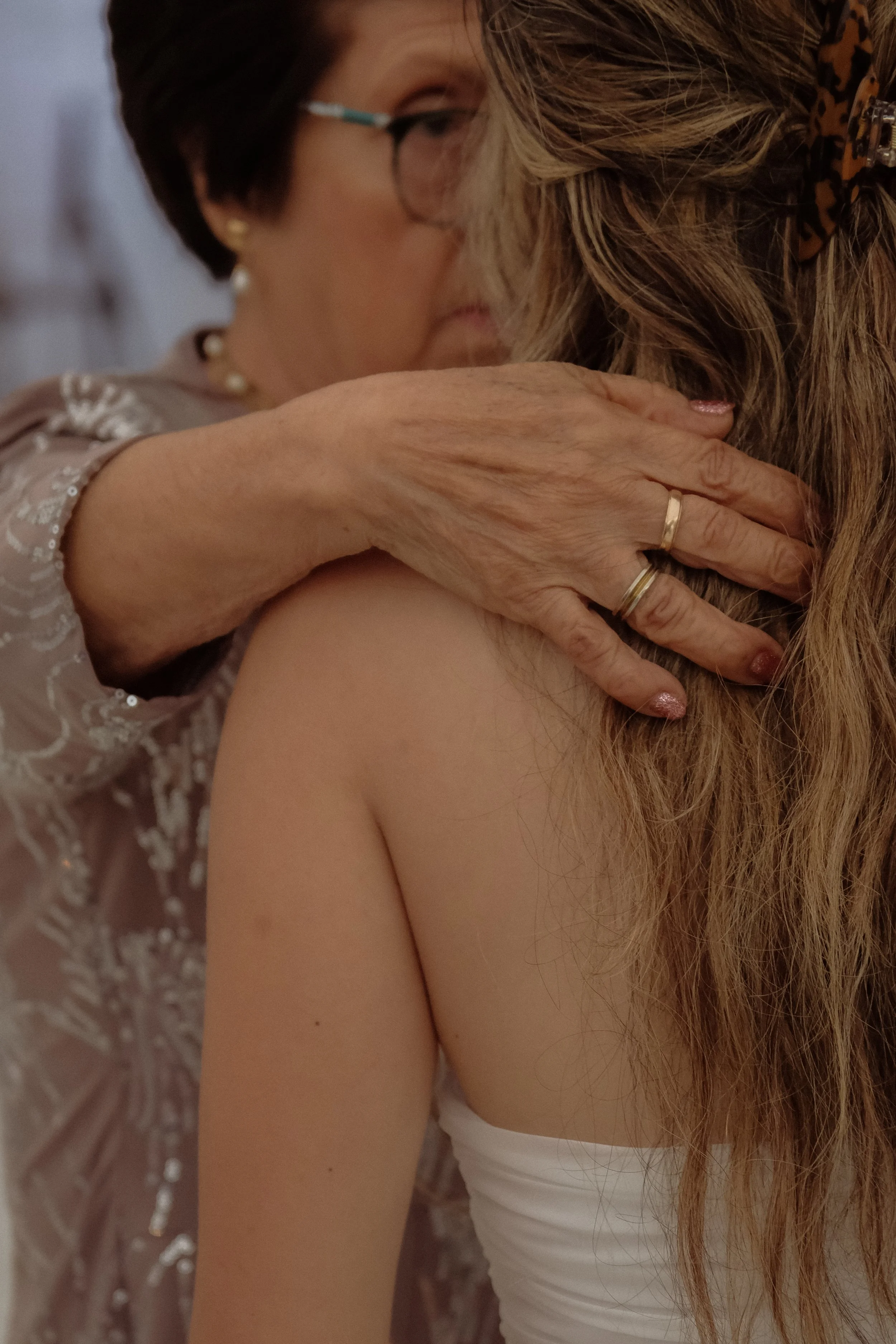 A close-up photograph of an elderly woman embracing a younger woman, both with their eyes closed. The elderly woman has short, dark hair, glasses, and is wearing a lacy patterned top, while the younger woman has long, light brown hair and is wearing 