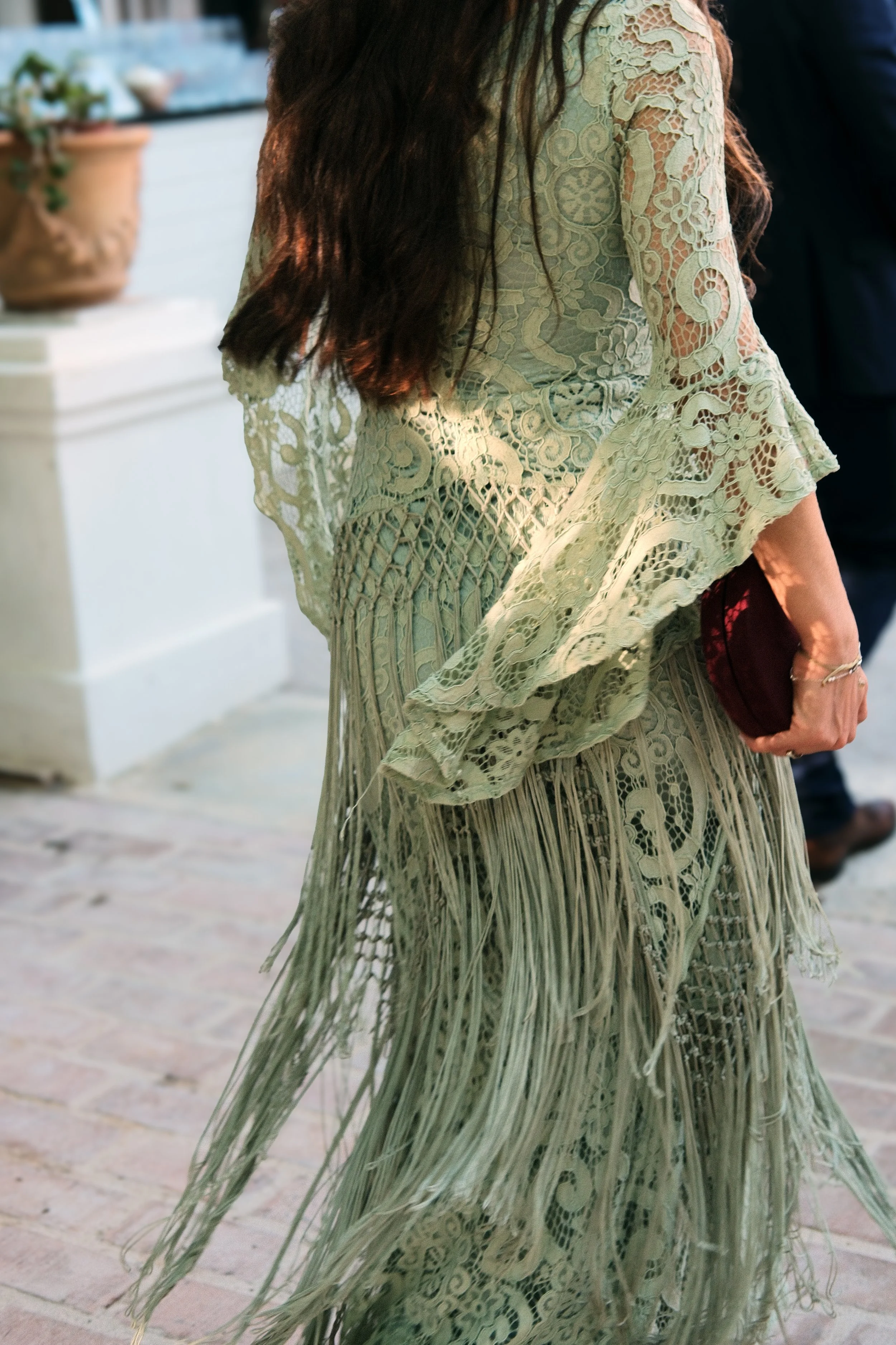 A woman with long dark hair wearing a green lace dress with intricate patterns and fringe details, holding a maroon clutch bag.