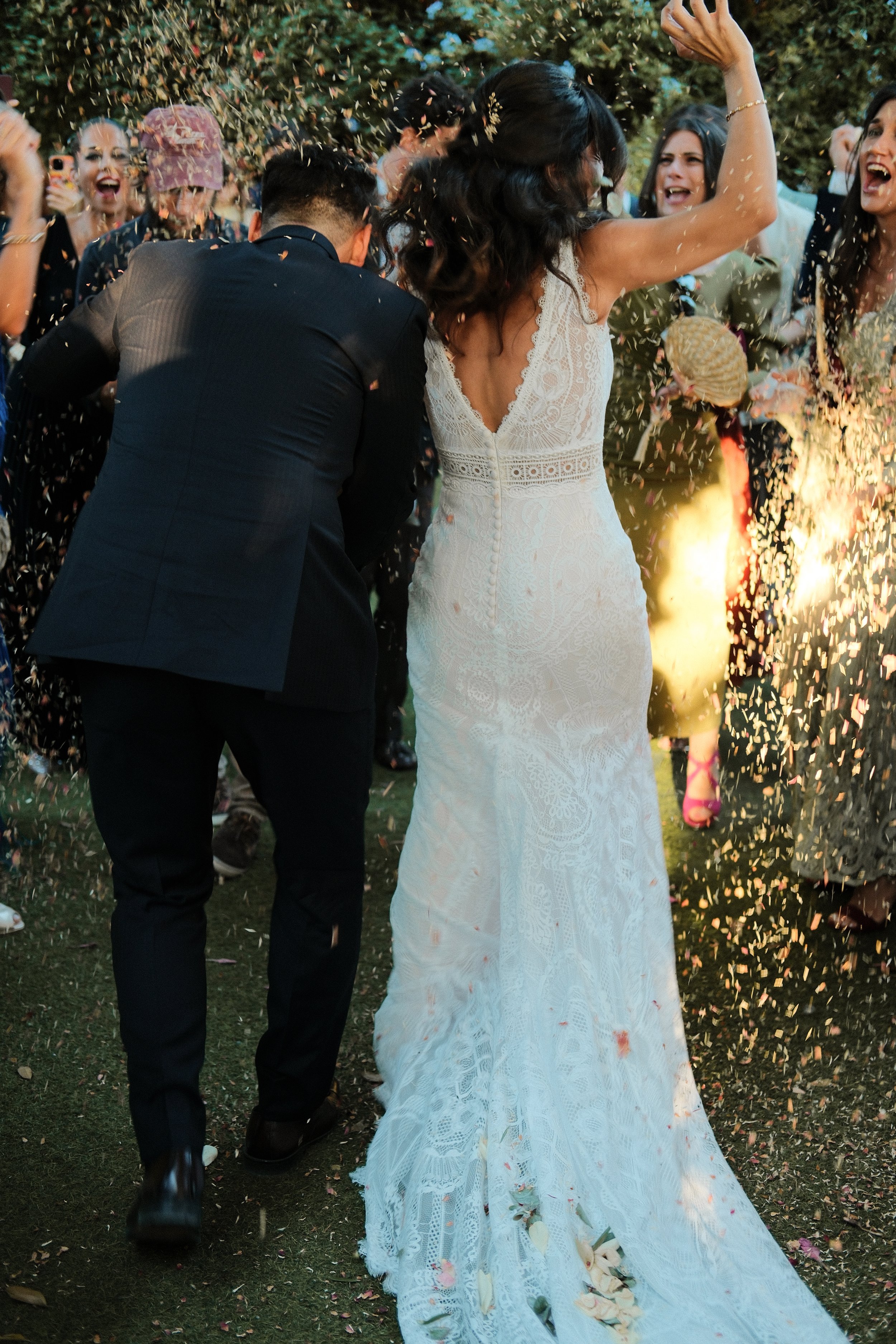 Bride wearing white lace wedding dress dancing outdoors with guests, confetti falling, and flames in background.
