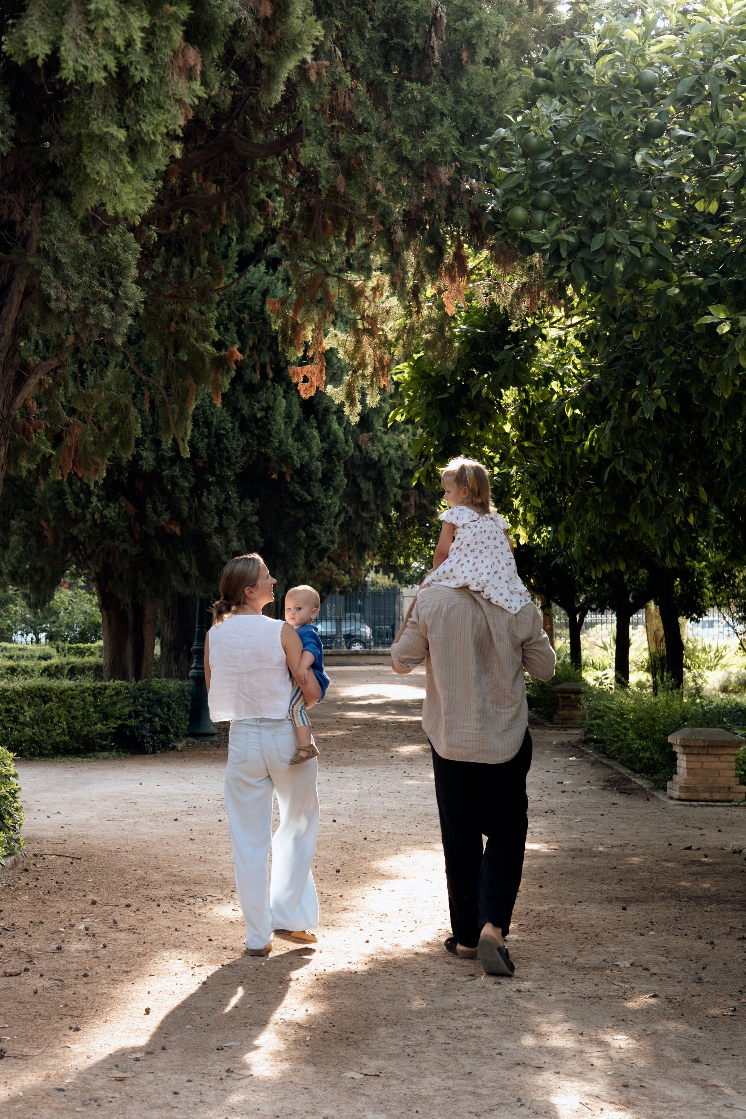 A woman carrying a child on her shoulders, a woman holding a young boy, and two children walking down a dirt path in a park with tall green trees and bushes.