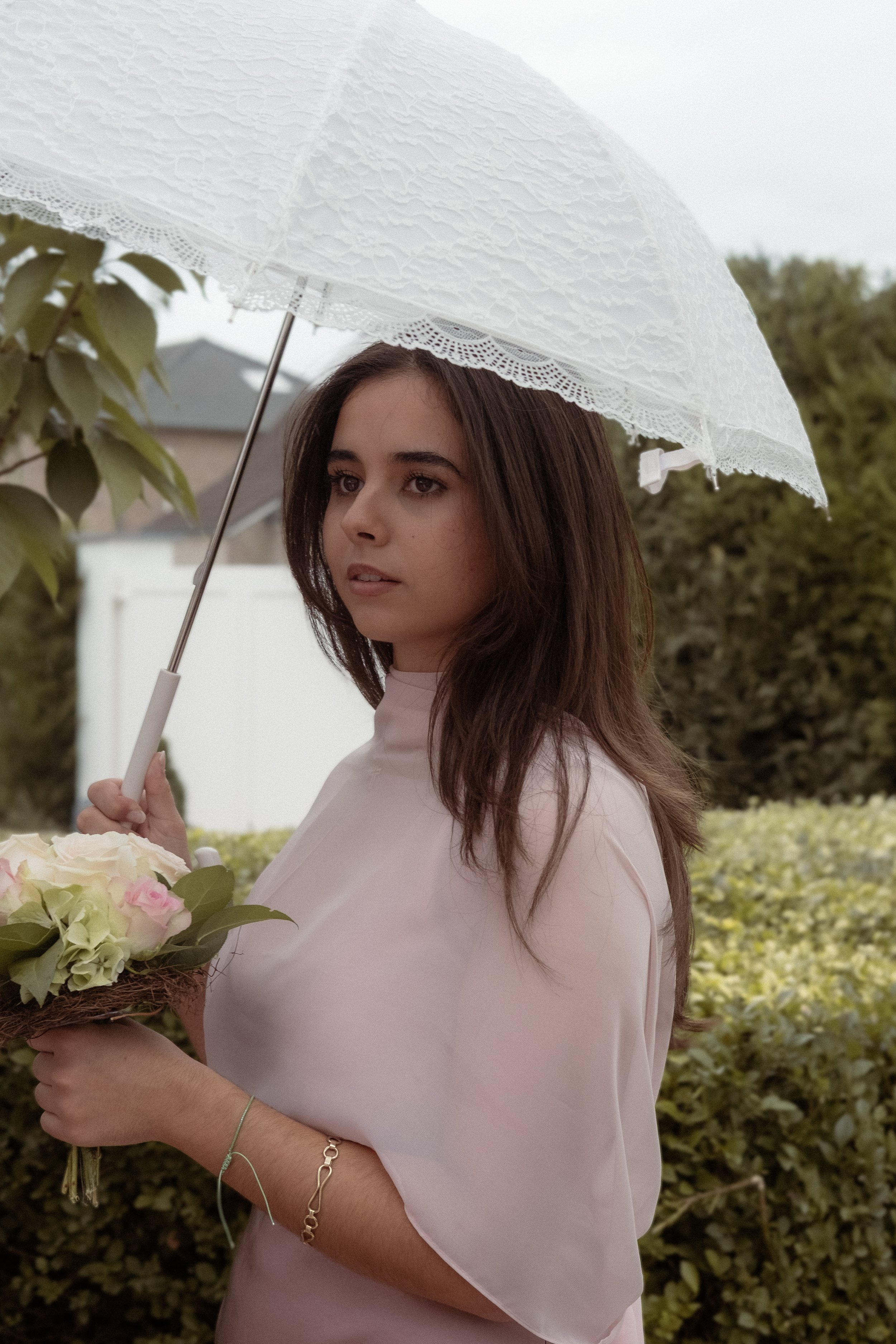 A young woman holding a flower bouquet with pink and white roses and greenery, standing outdoors with a white lace parasol, wearing a light pink dress, with trees and a fence in the background.