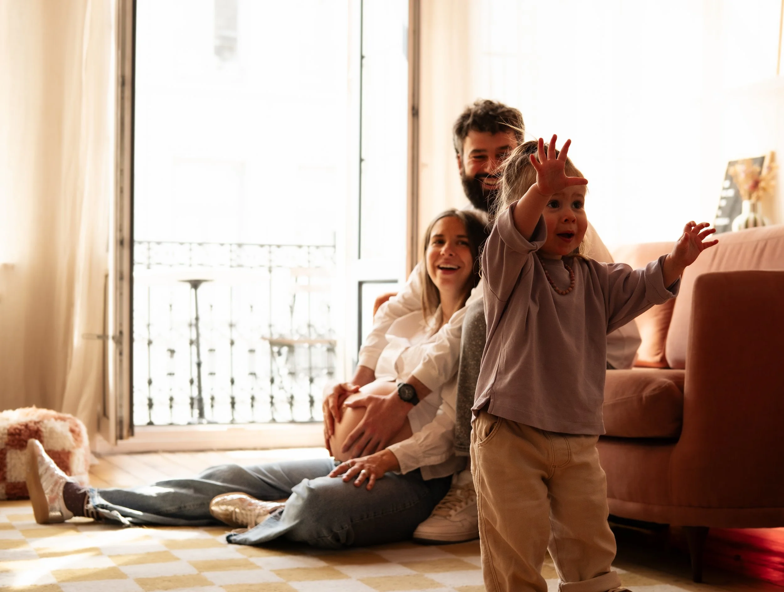 A young girl with blonde hair reaches out in front of a man and woman sitting on the floor of a sunlit living room. The man has a beard and is wearing a white shirt, and the woman has long brown hair. They are smiling and looking at the girl. The roo