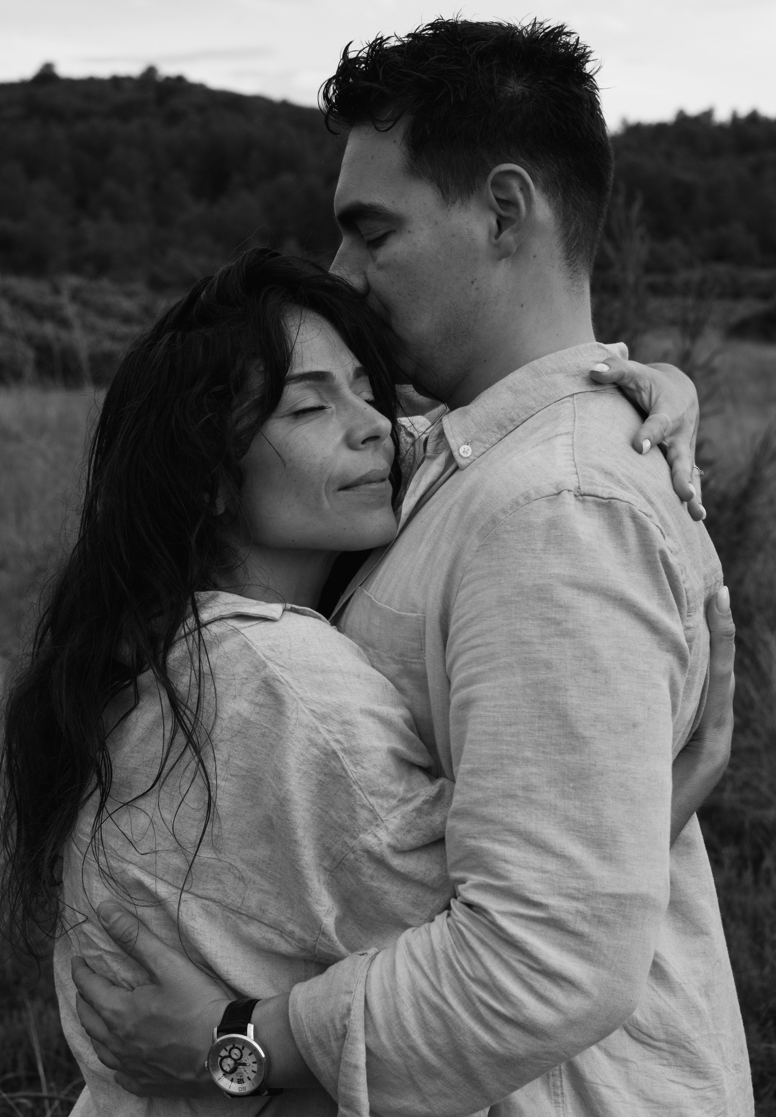 A black and white photo of a couple hugging outdoors, with the woman smiling with eyes closed and the man kissing her on the forehead.