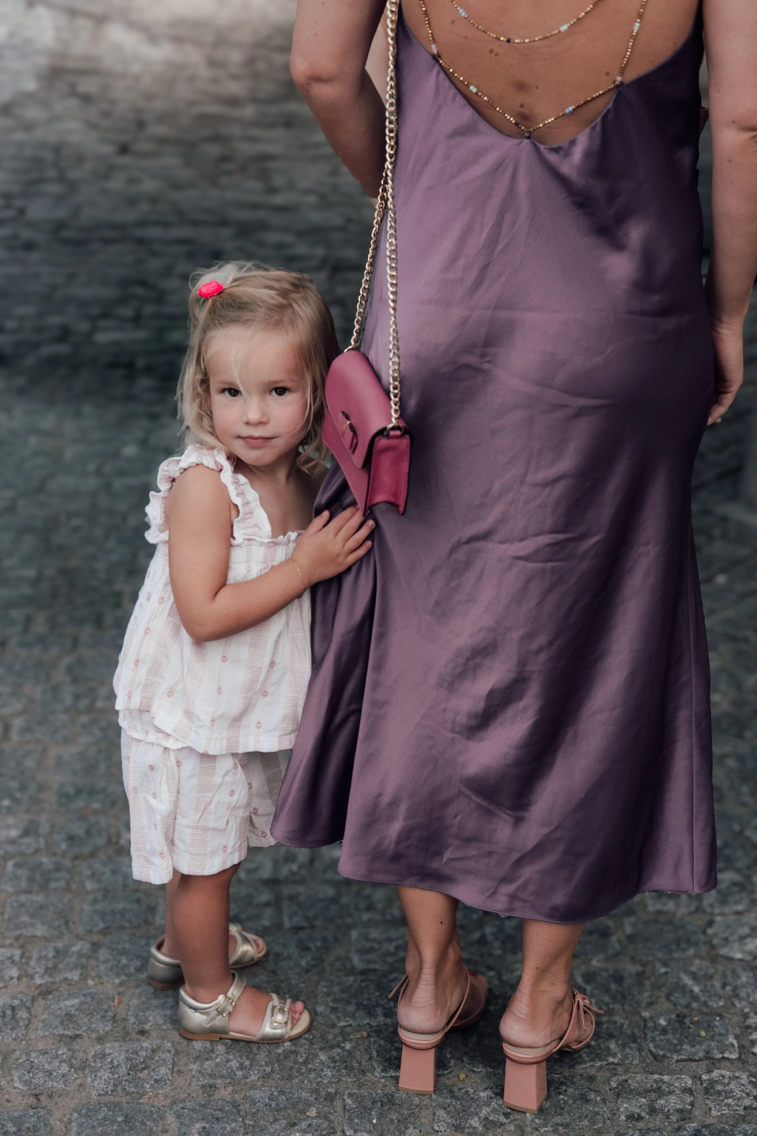 A young girl with blonde hair, wearing a white striped dress, standing on cobblestone pavement and holding onto an adult's dress. The adult, wearing pink high-heeled shoes, purple satin dress, and gold jewelry, is partially visible. The background sh