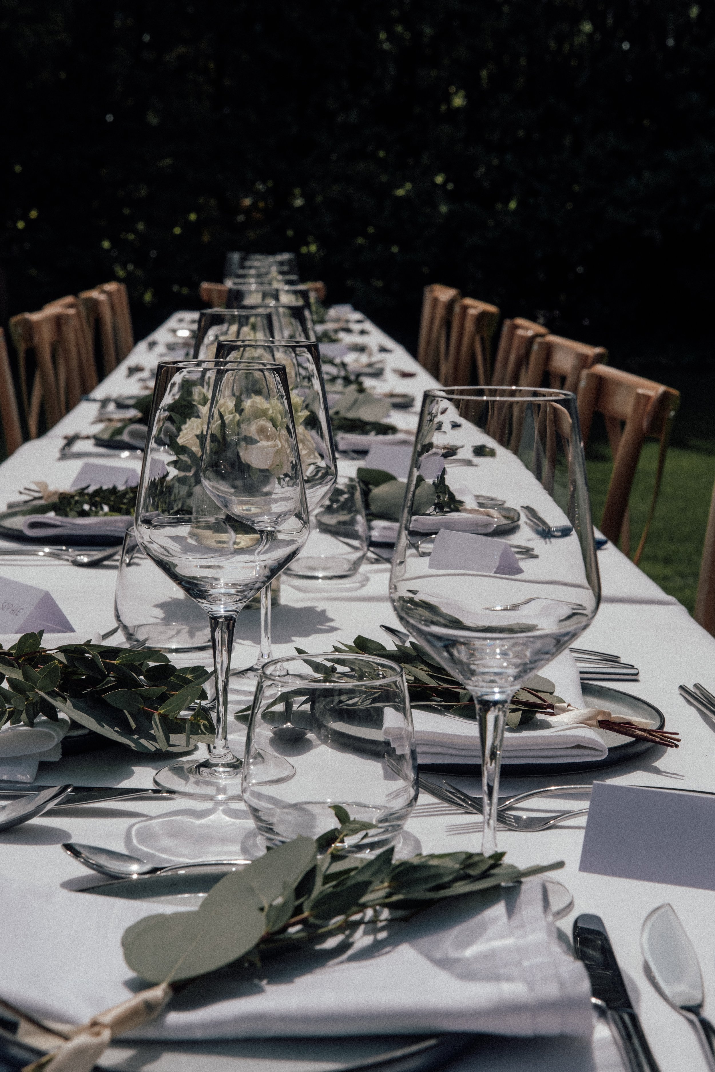 Long outdoor dining table set for a formal event with wine glasses, white napkins, and greenery decorations, with wooden chairs and a dark outdoor background.