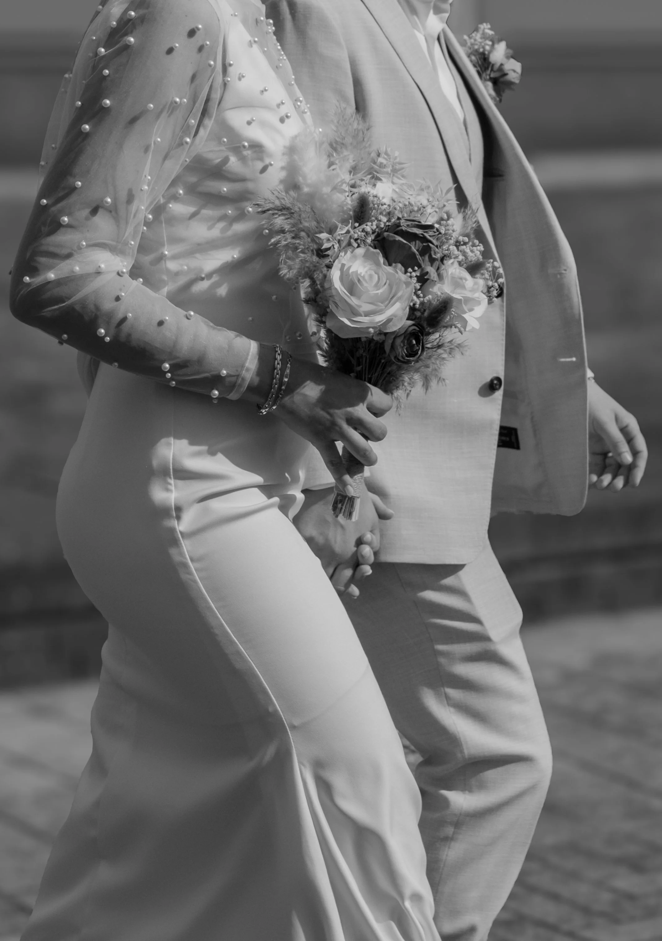 A bride and groom holding hands, the bride holding a bouquet of flowers, wearing a dress with pearl embellishments on the sleeves, and the groom in a suit, walking together outside.