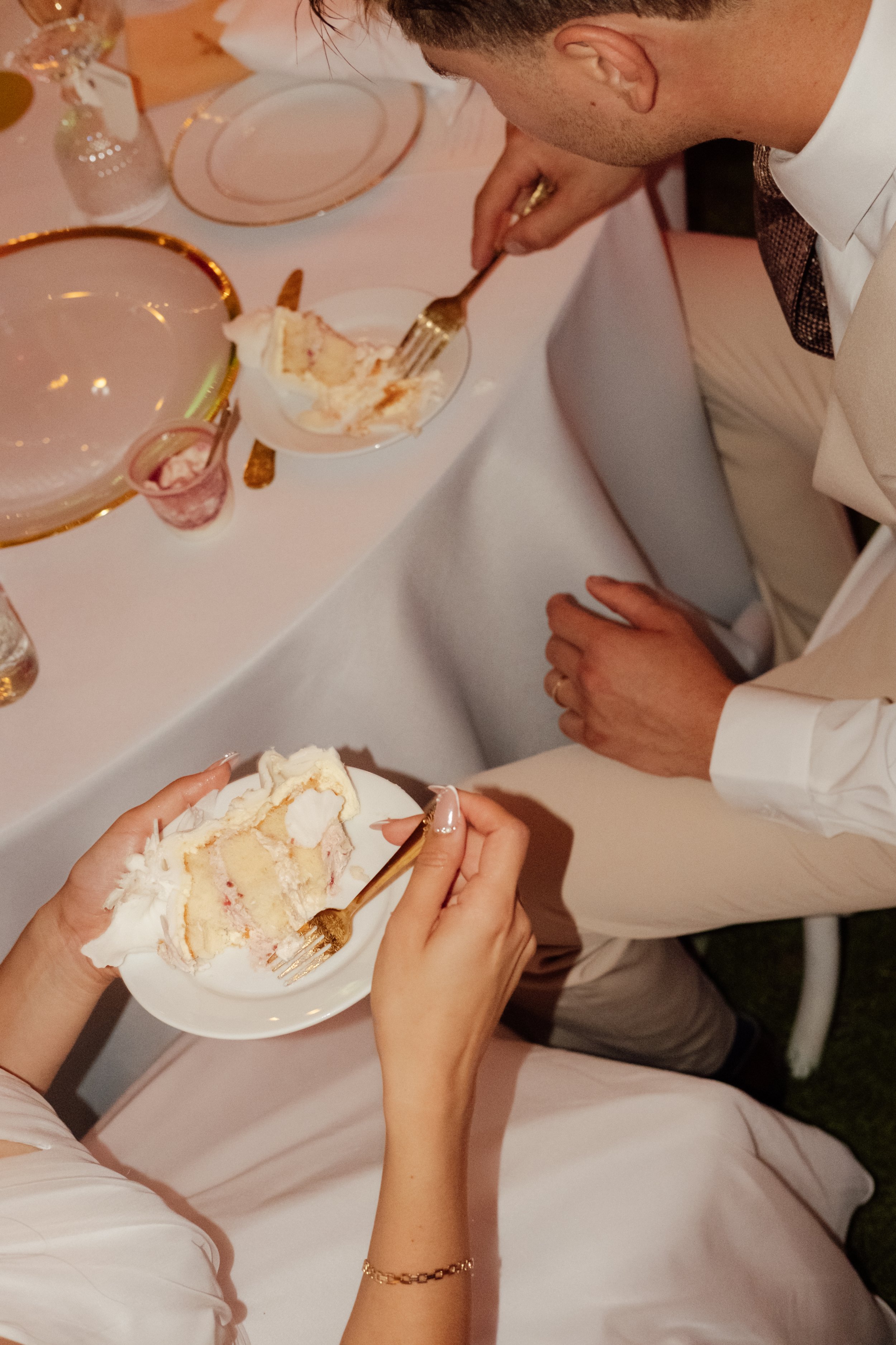 People at a celebration eating cake, with a focus on one person holding a plate with a slice of cake and a fork. The cake is light-colored with pink filling and white icing. The table is decorated with plates, glasses, and gold trim.