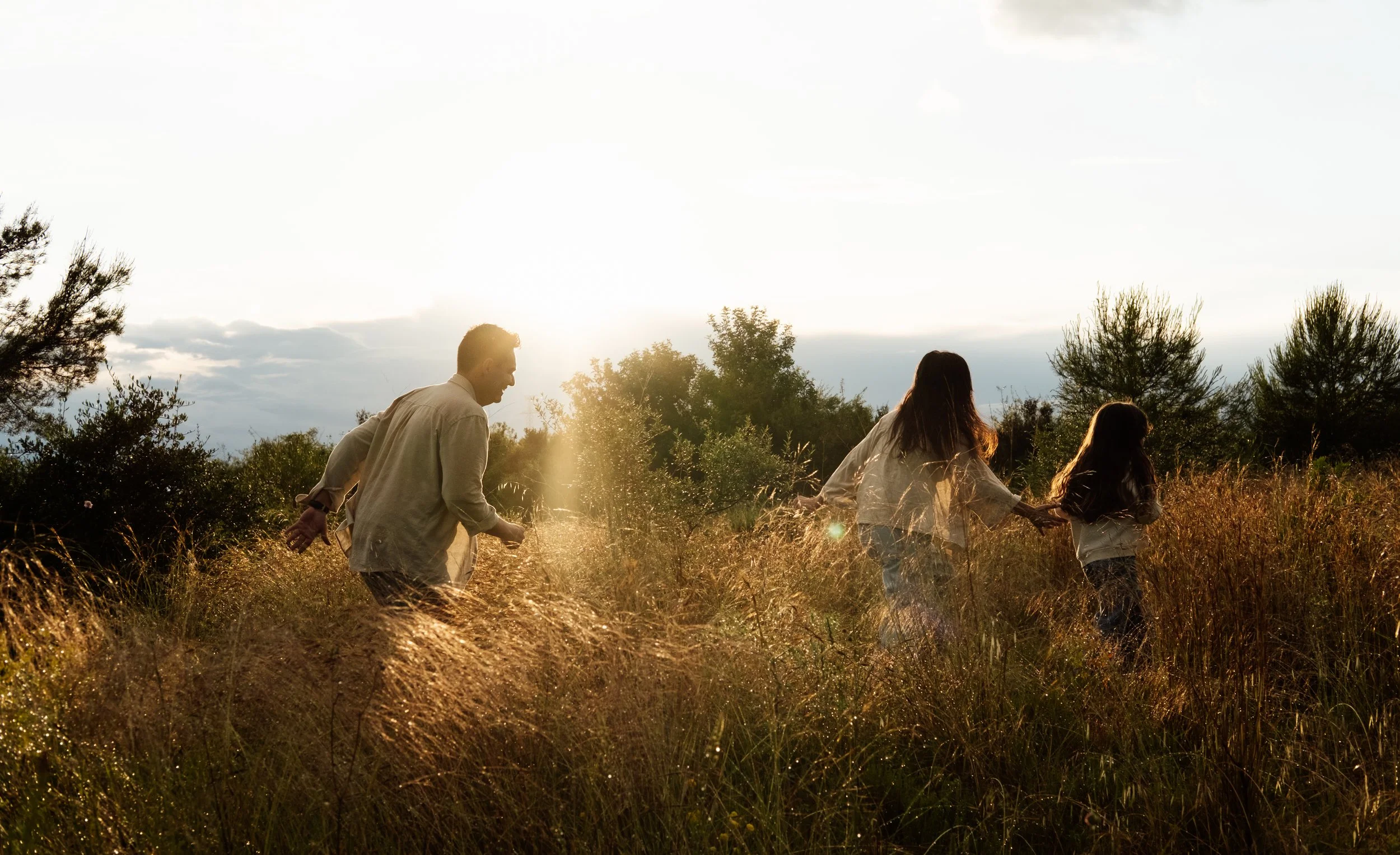 Three people, two women and one man, walking hand in hand through a golden grassy field during sunset, surrounded by trees.
