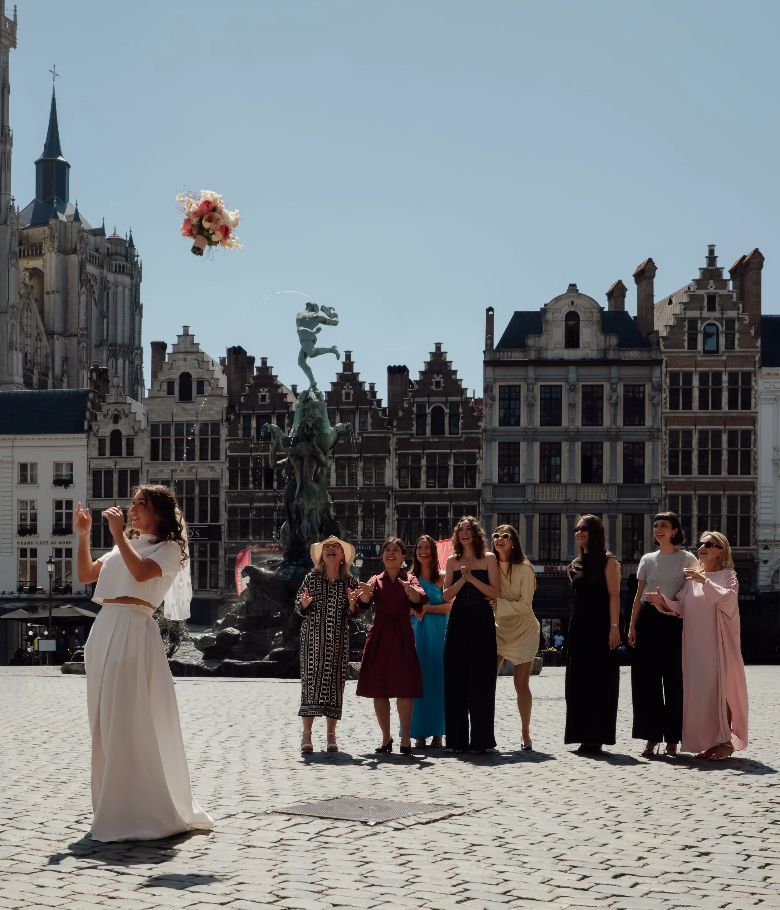 Women in elegant dresses standing in a historic European city square, some laughing and smiling, with a fountain and iconic Gothic-style buildings in the background.