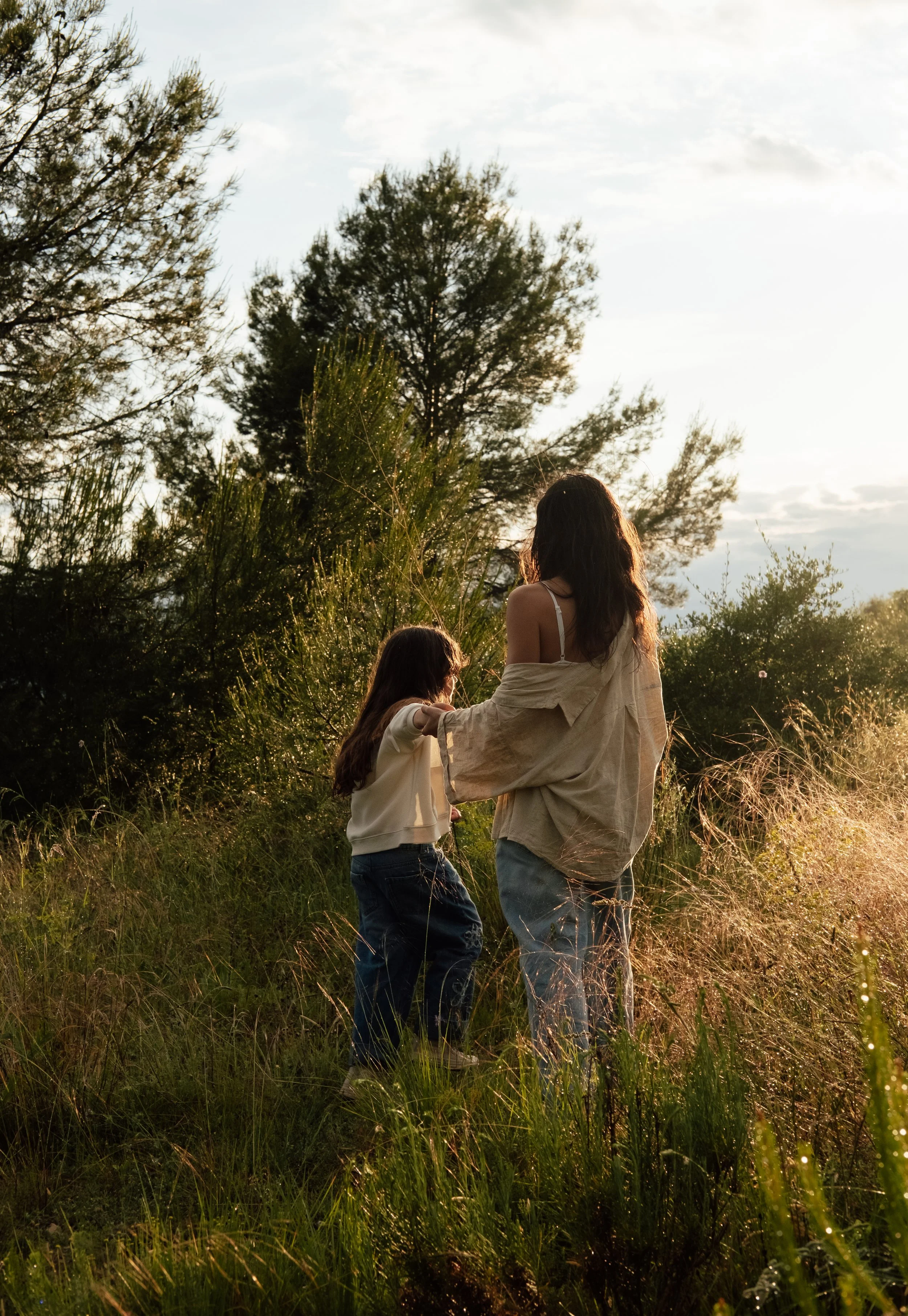 A woman and a girl stand in a grassy field with tall trees in the background, illuminated by golden sunlight.