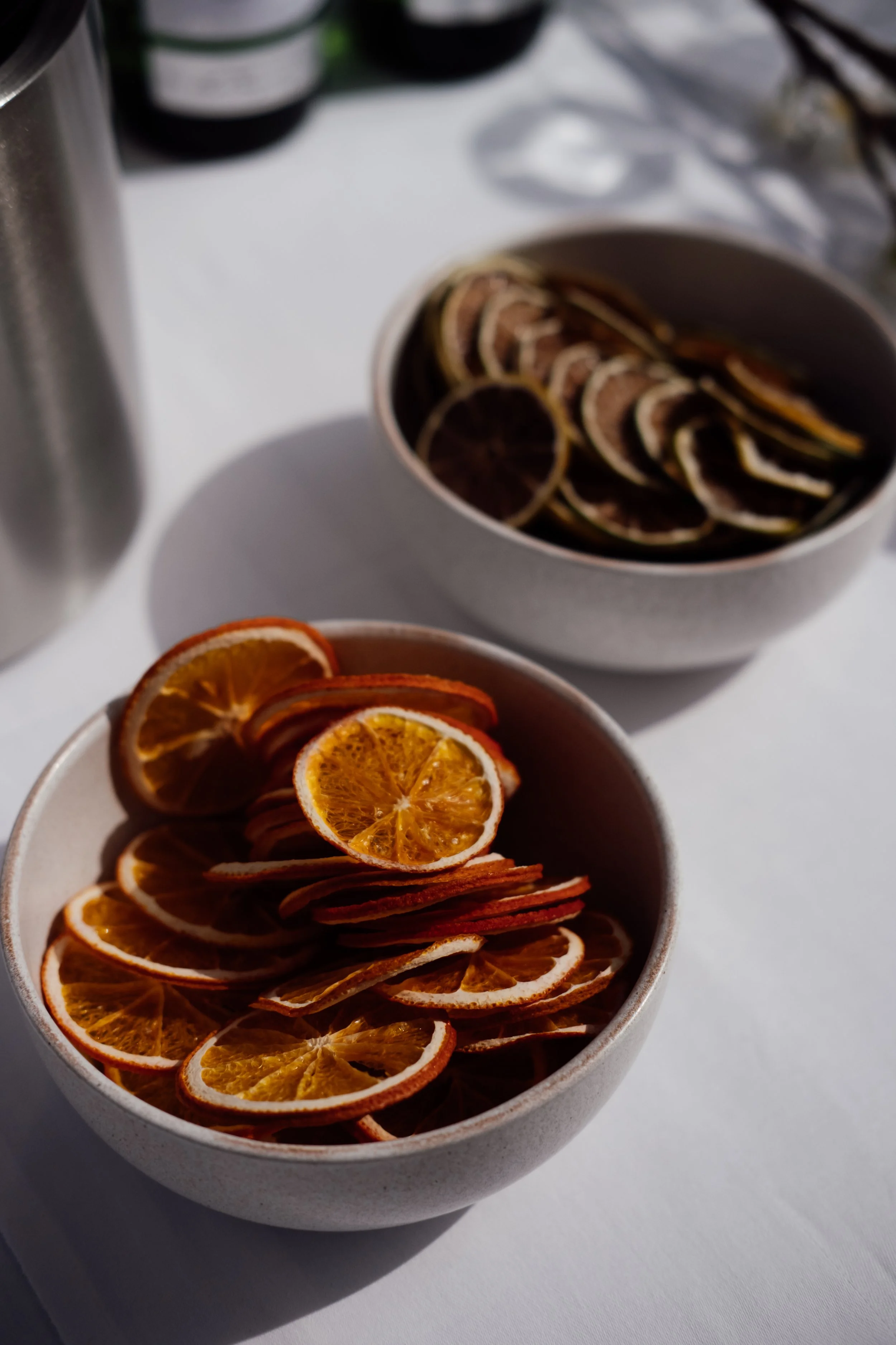 Close-up of dried orange slices in a white bowl with a shadow cast on a white surface, with an out-of-focus container and dark bottles in the background.