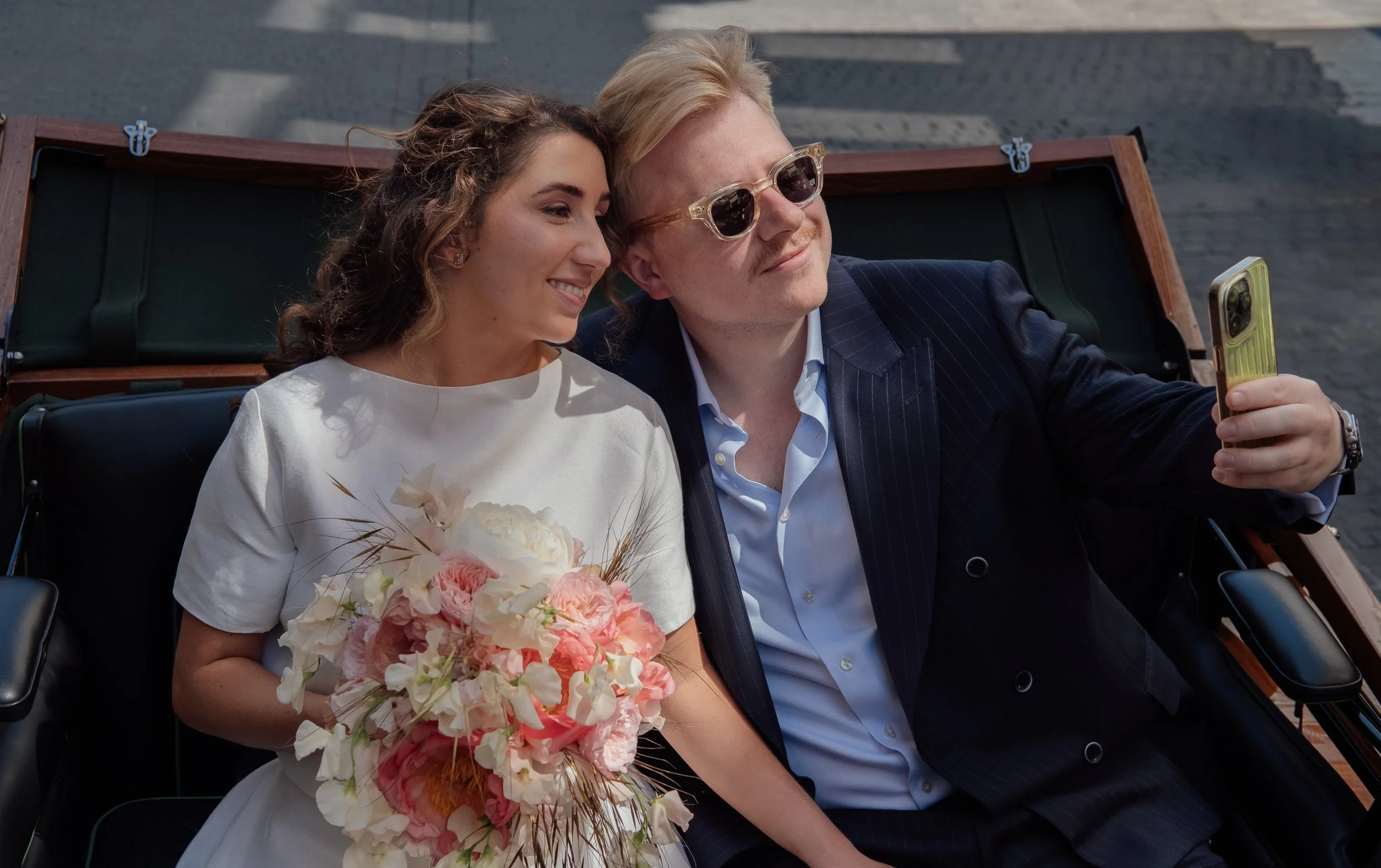 A woman in a white dress holding a bouquet of pink and white flowers, sitting next to a man in sunglasses and a navy blazer, both taking a selfie together on a boat.