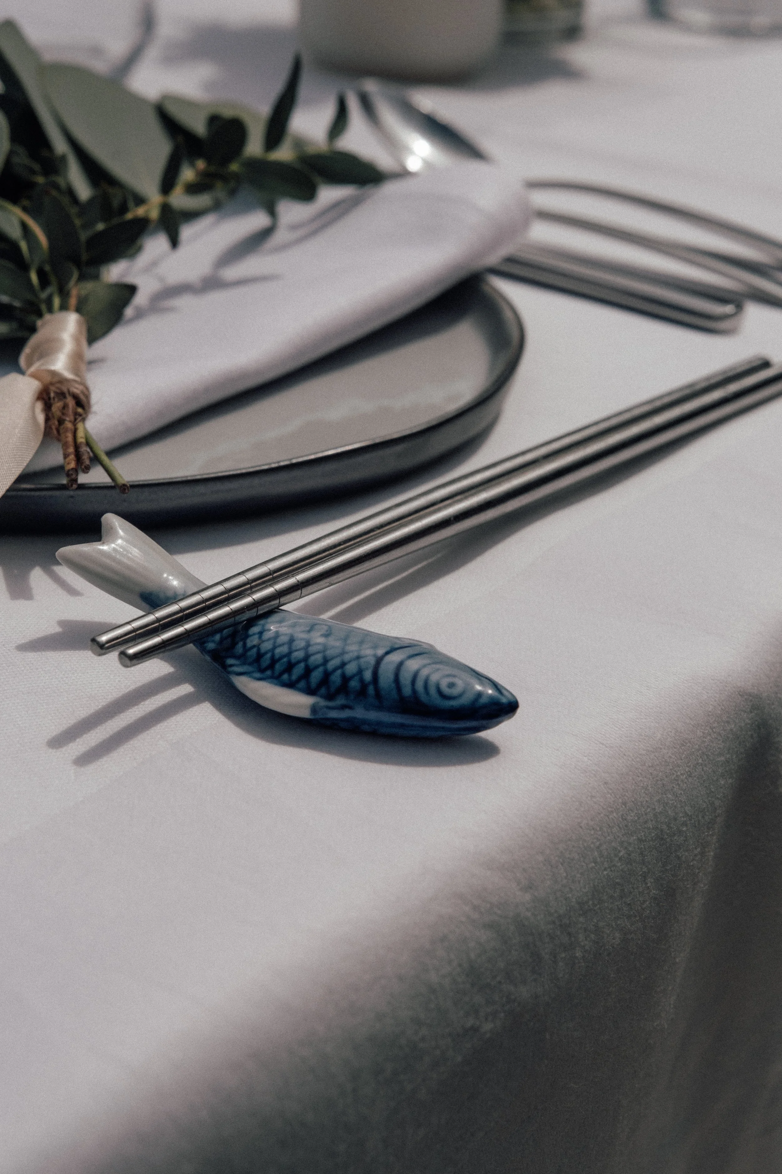A place setting with a fish-shaped chopstick rest, silver chopsticks, a silver spoon, a white napkin, and a leafy branch on a white tablecloth.
