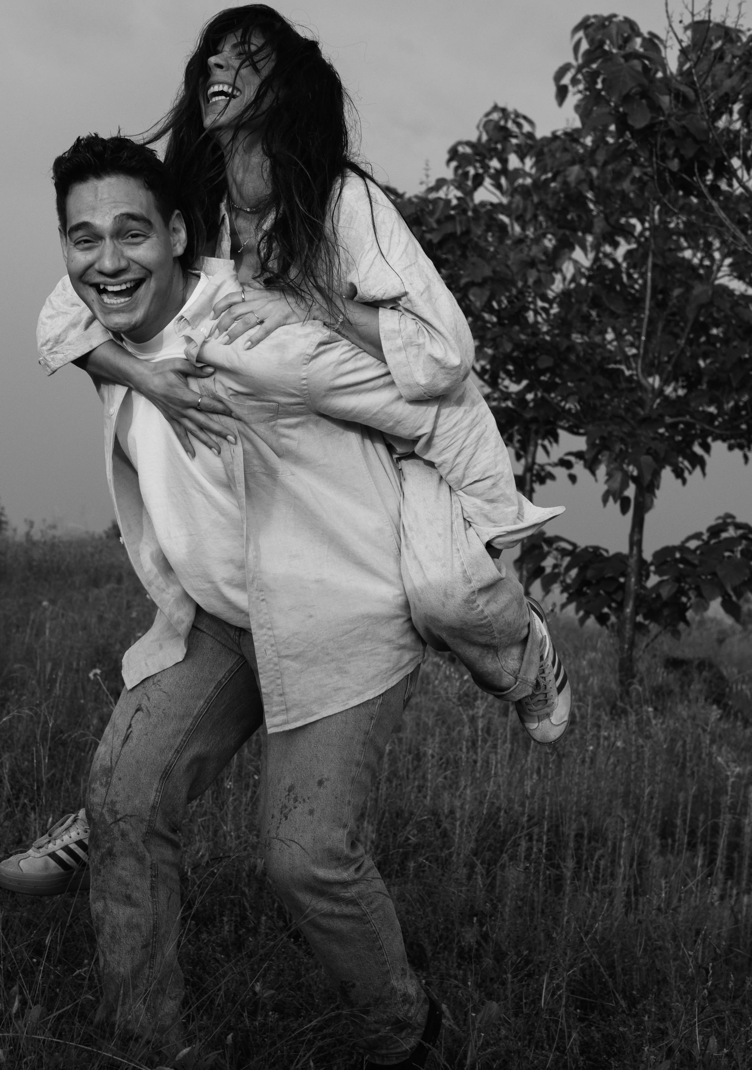 A black and white photo of a man giving a piggyback ride to a woman outdoors on a grassy field with trees in the background, both smiling and appearing happy.