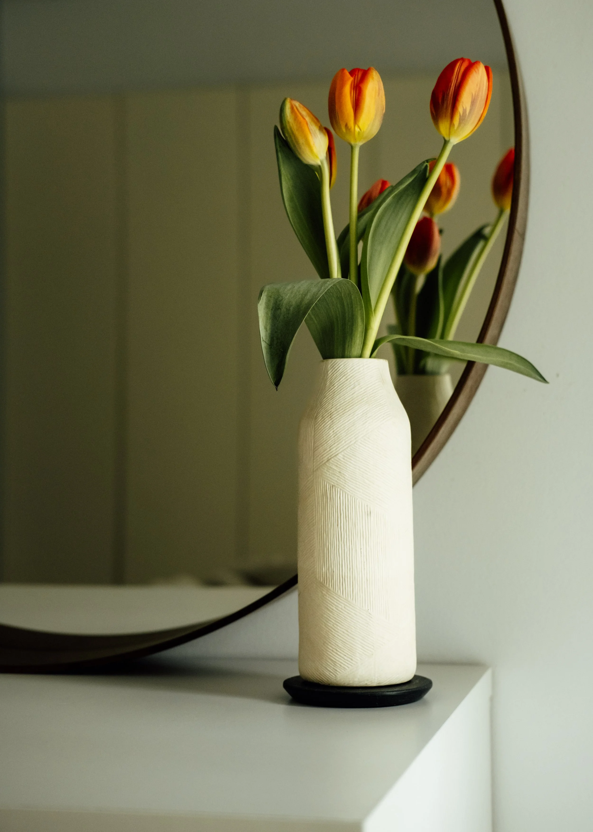 A white textured ceramic vase with a leaf pattern, holding a bouquet of orange and yellow tulips, reflected in a round mirror behind it, on a white surface.