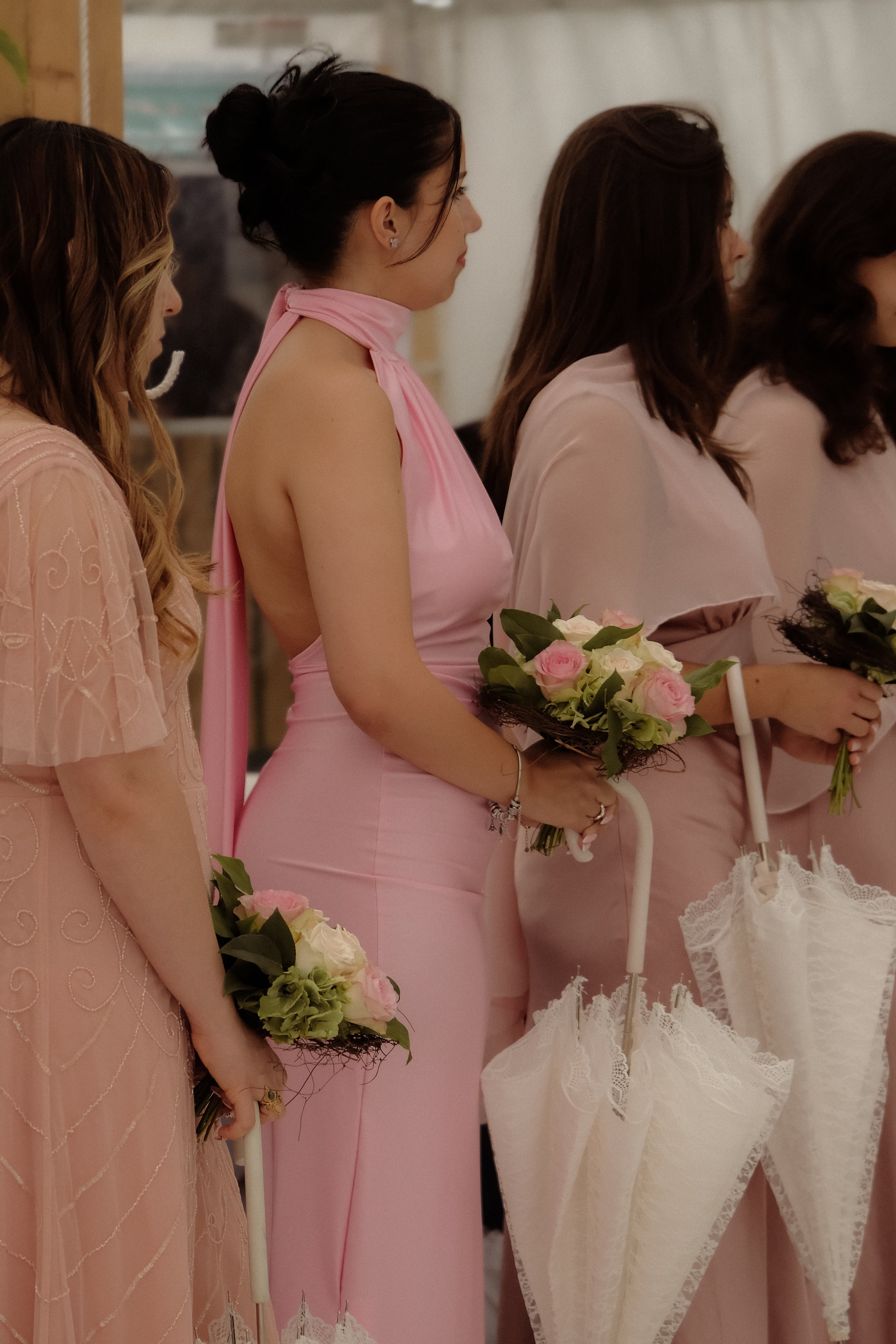 Line of women dressed in soft pink and beige dresses holding flower bouquets, standing next to each other at a wedding or formal event.