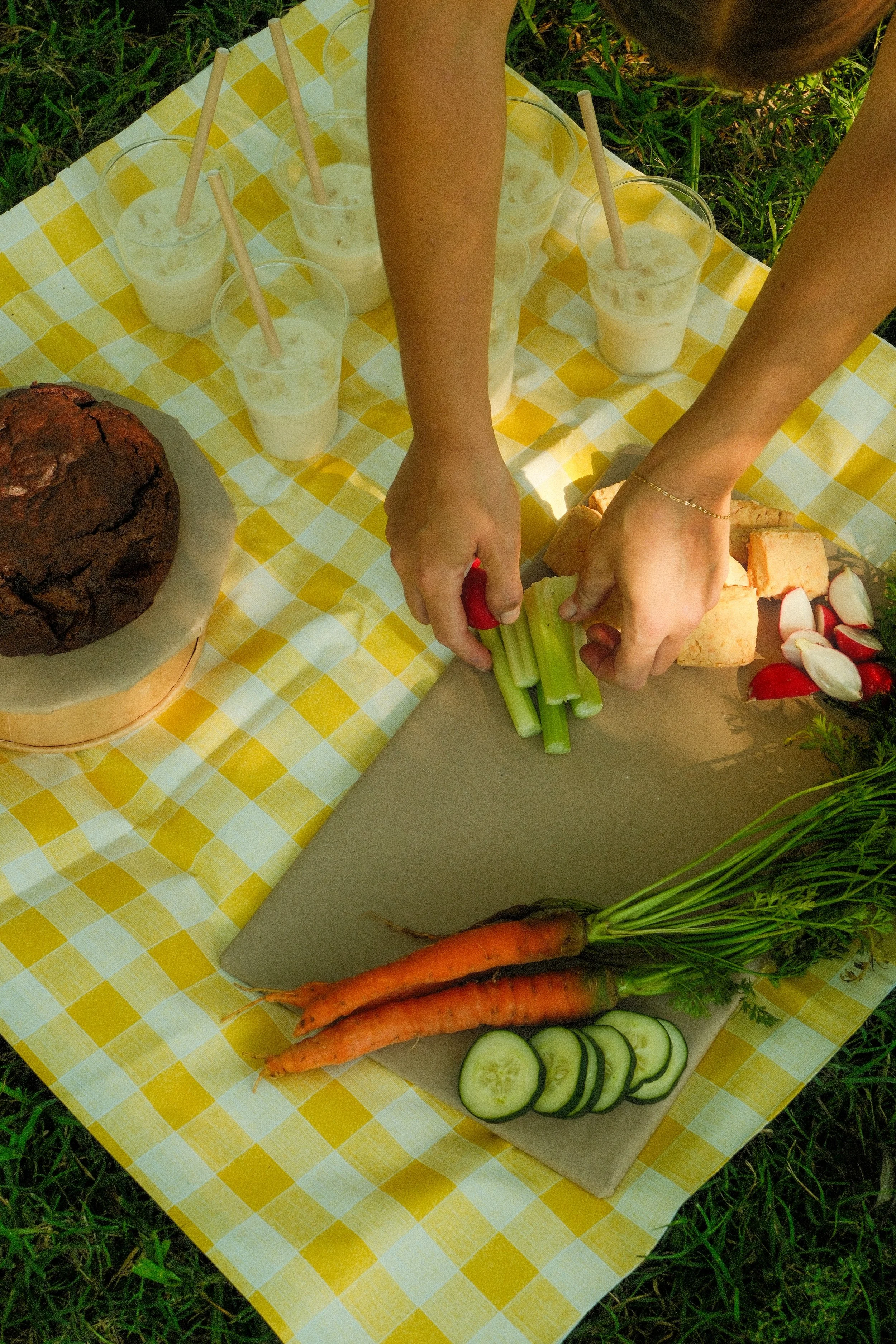 Person slicing celery on a cutting board surrounded by carrots, radishes, and bread, with glasses of milk and a chocolate cake on a yellow checkered tablecloth outdoors.