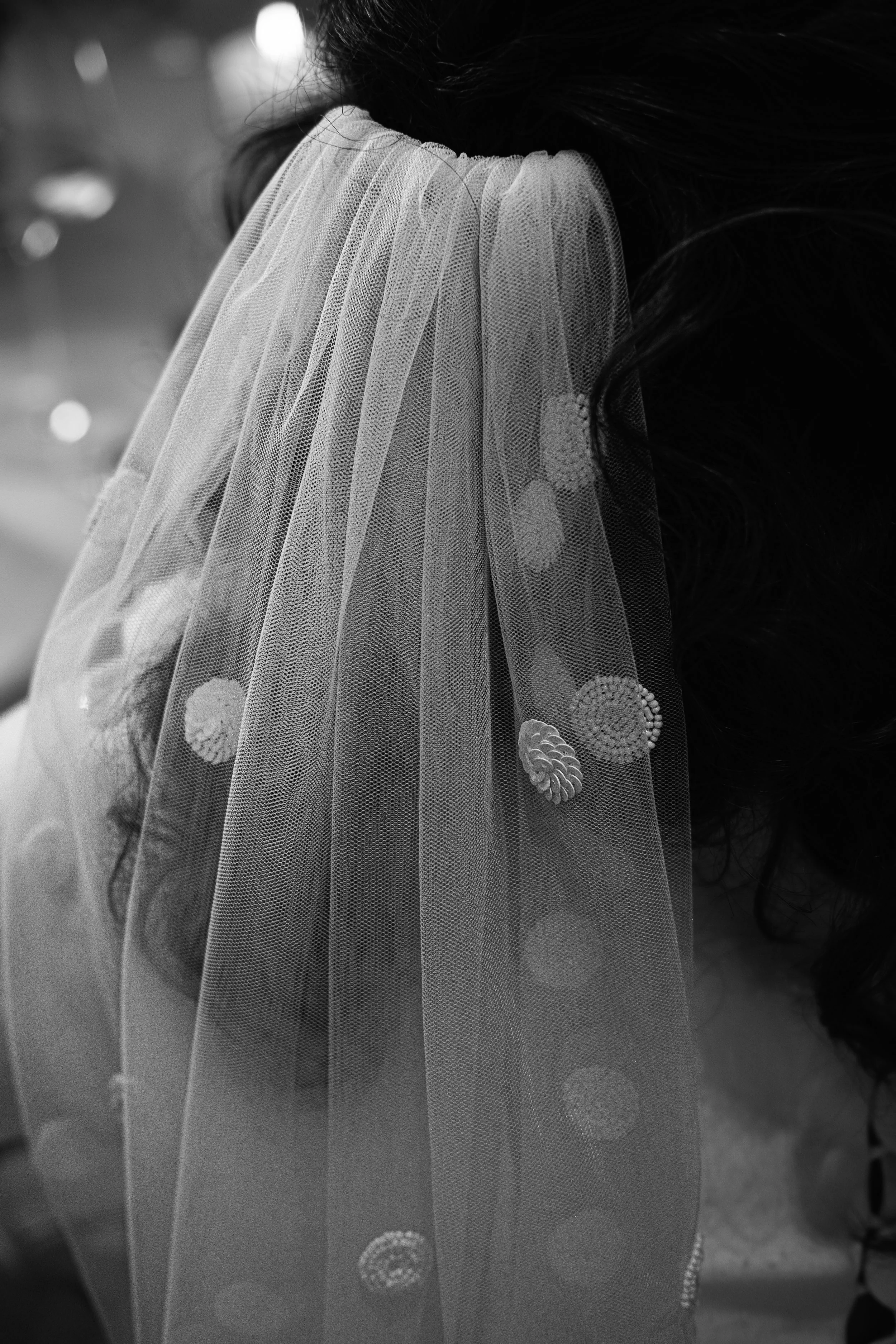 Close-up black and white photo of a woman's shoulder, partially covered by a sheer, embellished veil with circular patterns.