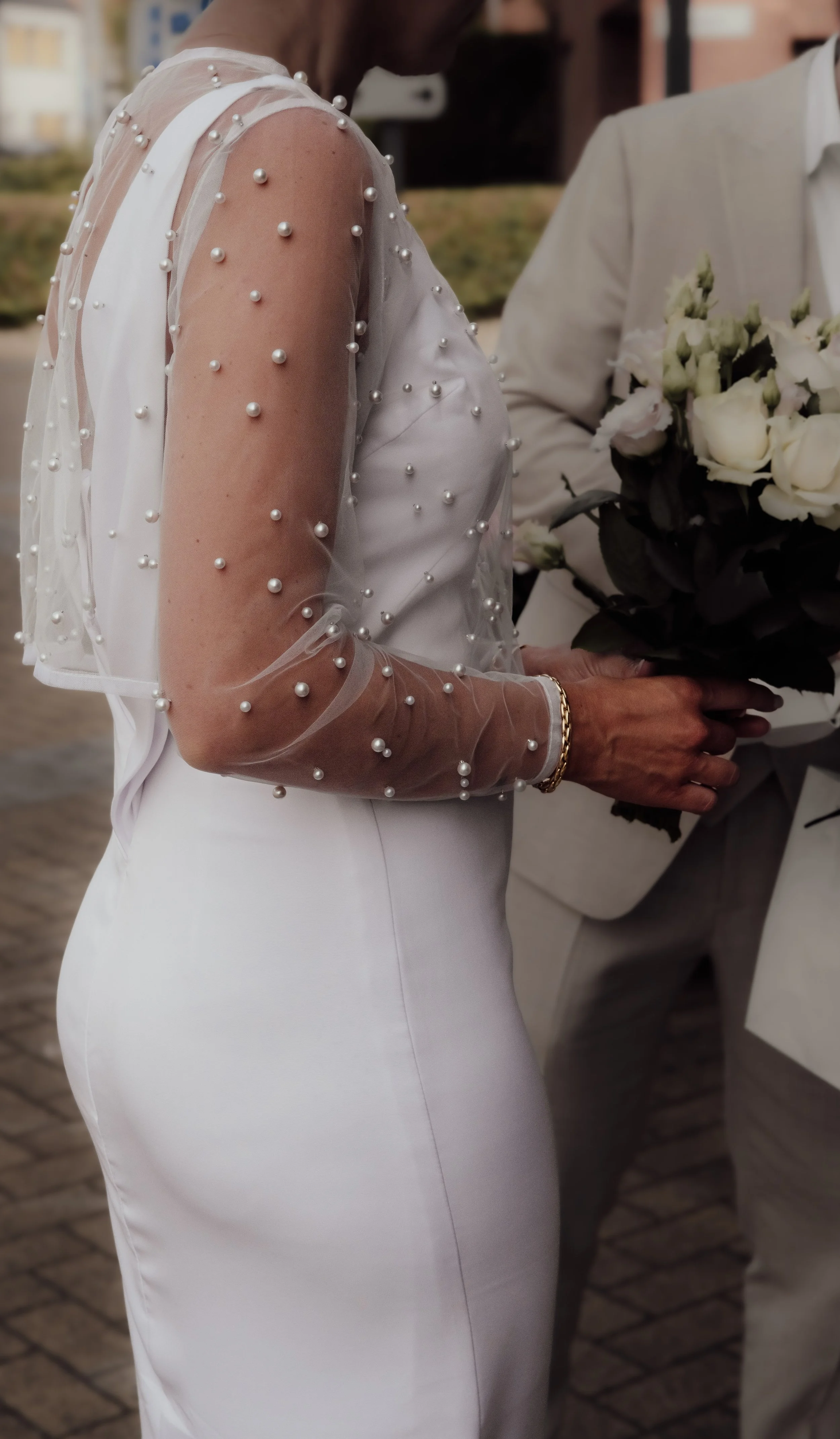 A woman in a white dress with sheer, pearl-embellished sleeves is holding a bouquet of white roses.
