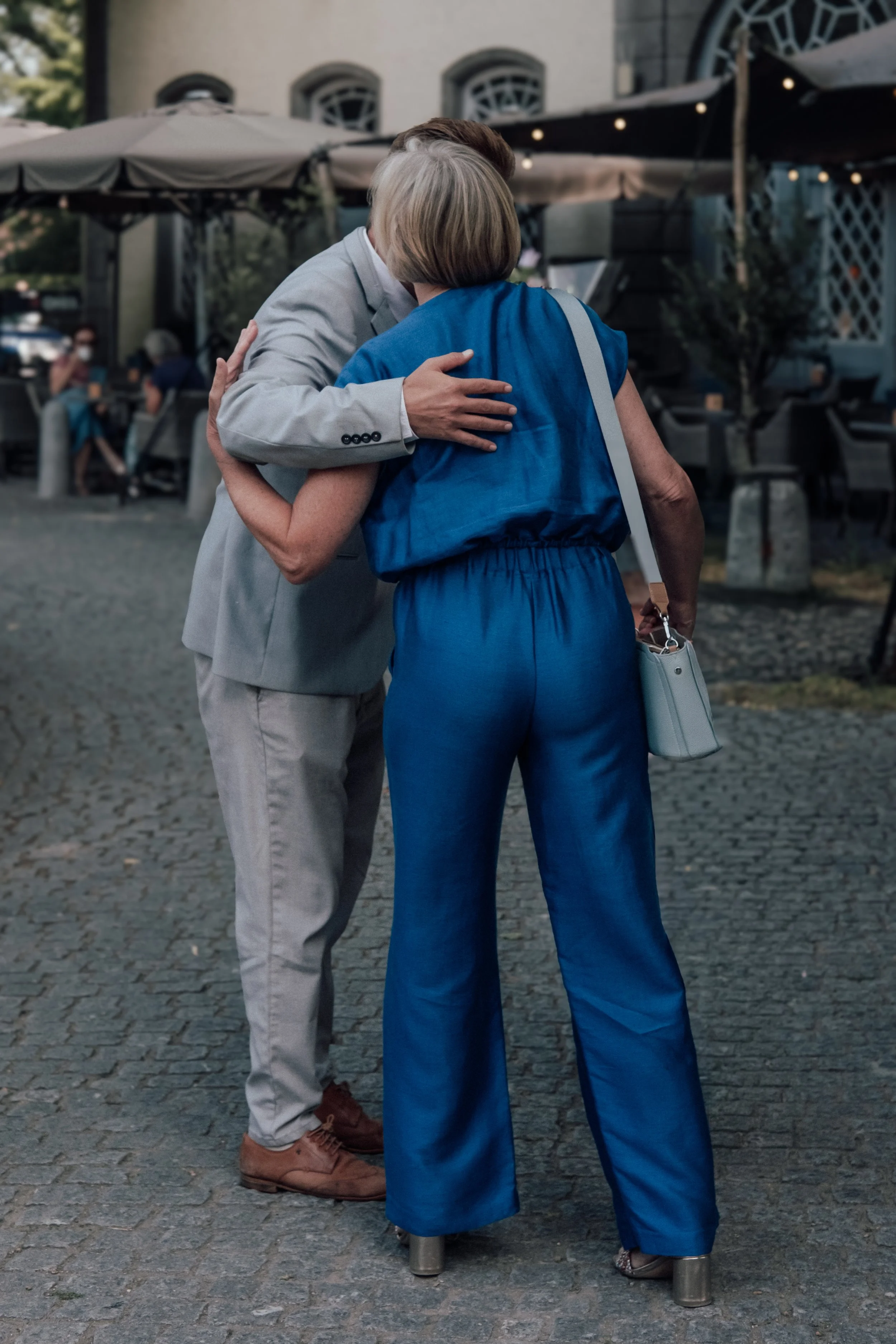 A man in a light gray suit hugging an elderly woman in blue clothing outside on a cobblestone street, with outdoor seating and a building in the background.