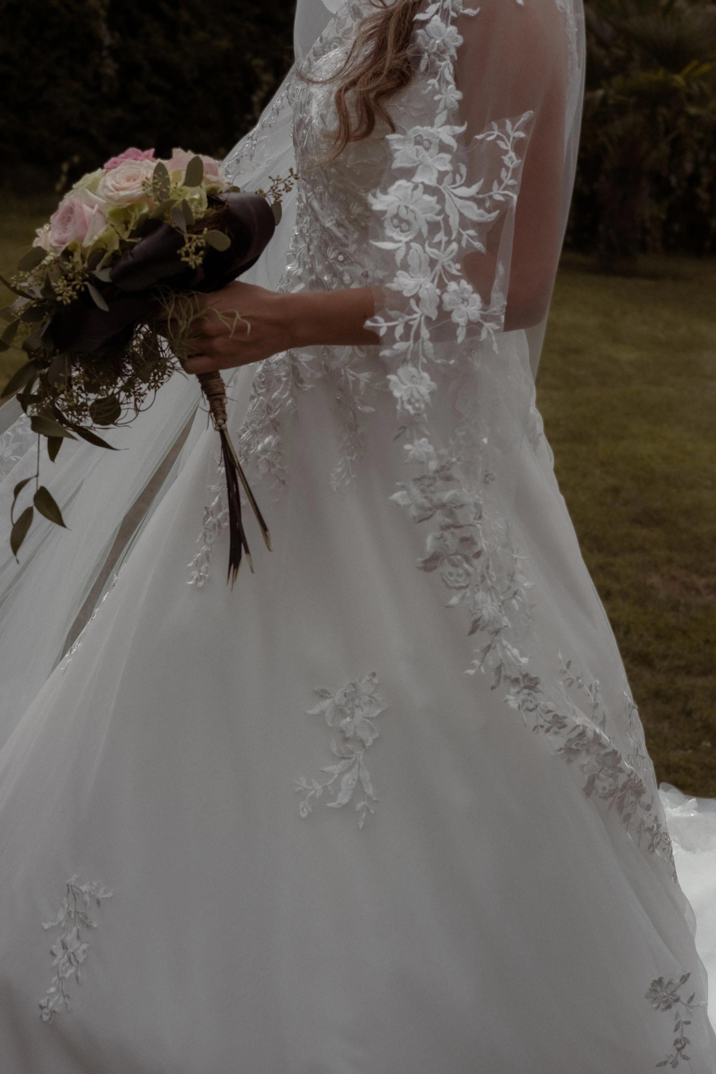 A bride holding a bouquet of pink and white roses with greenery, wearing a white lace wedding dress with floral embroidery, outdoors during the evening or nighttime.