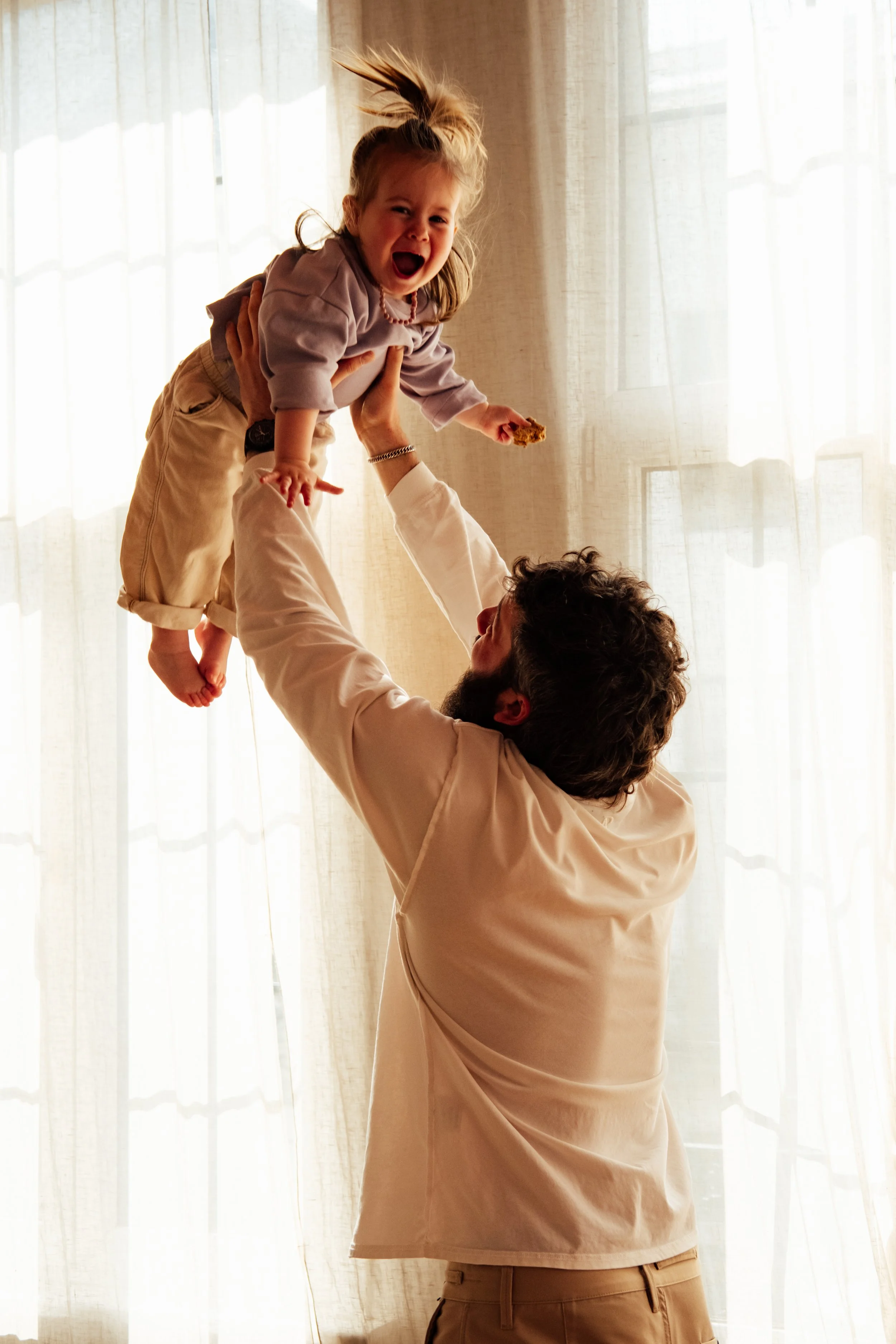 A man lifting a young girl with blonde hair into the air. Both are indoors in front of a sheer curtain, with natural light coming through.