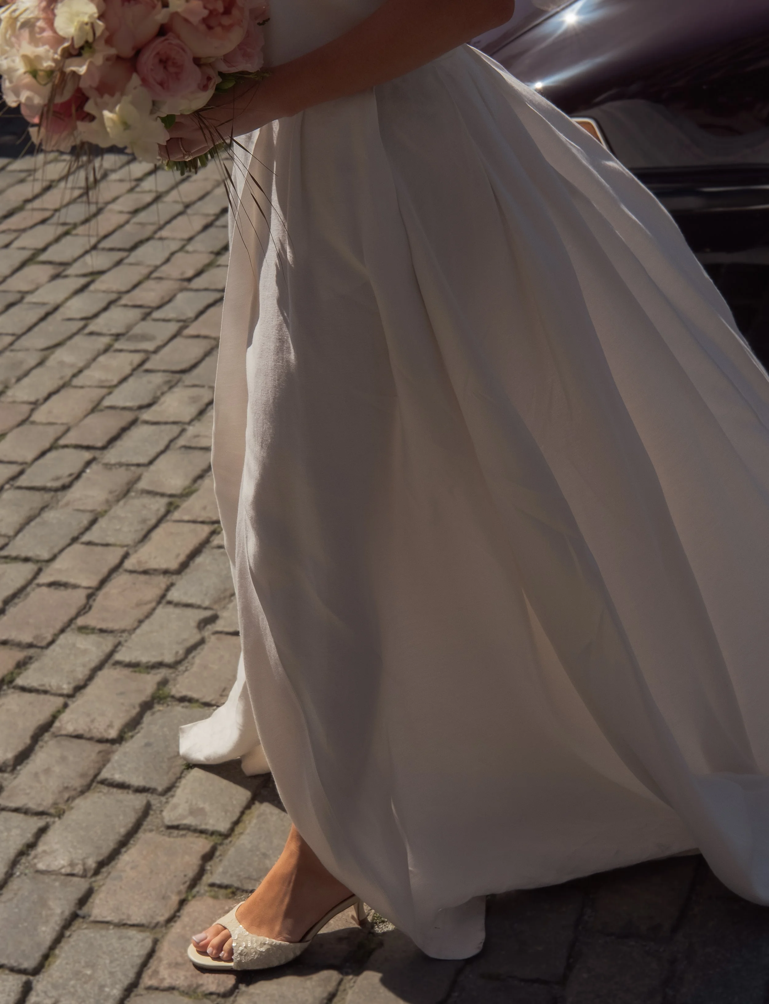 Close-up of a bride's hand holding a bouquet of pink and white flowers, wearing a white wedding gown, walking on cobblestone pavement.