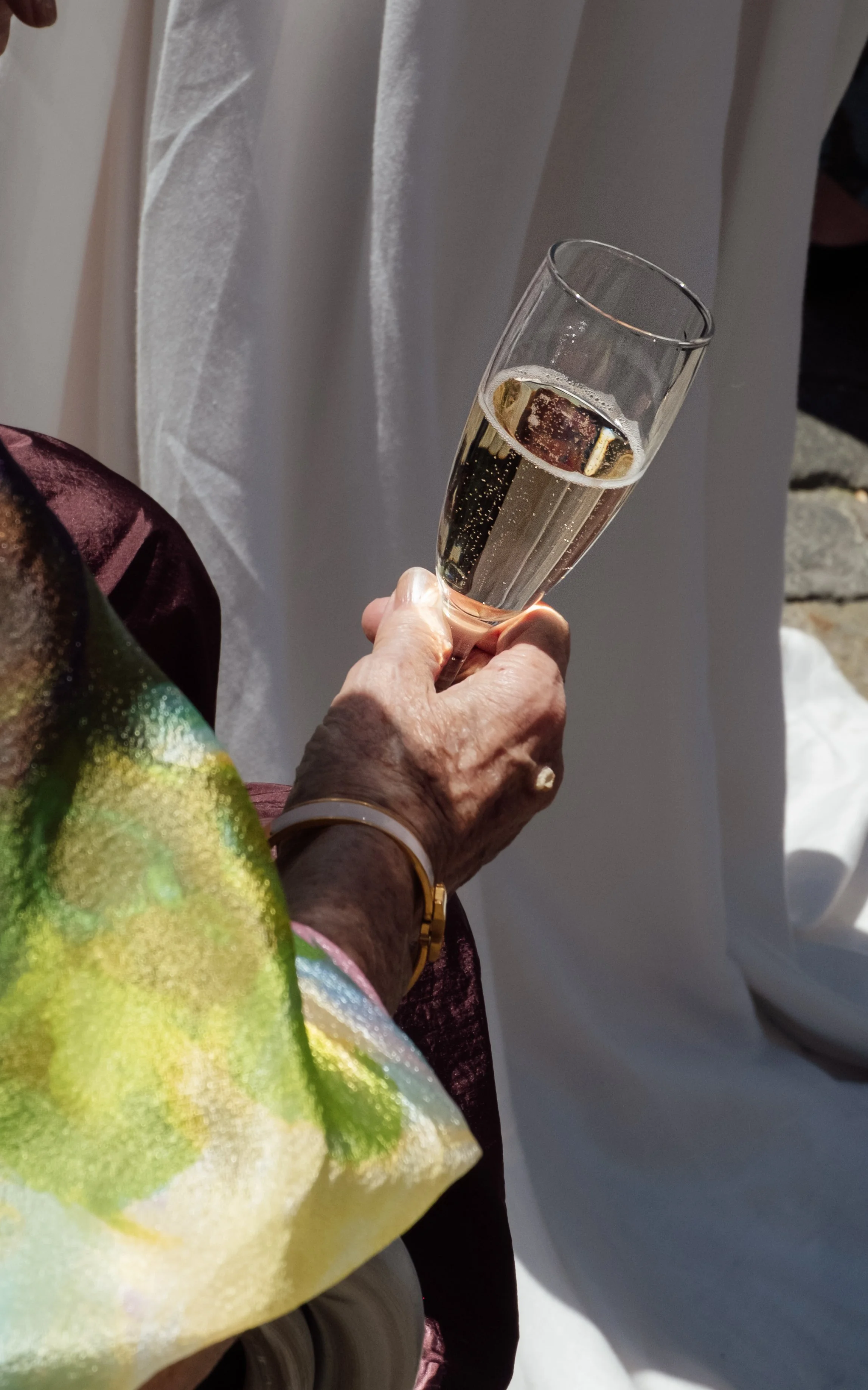 A person holding a champagne flute filled with sparkling wine or champagne, with a white curtain and bench visible in the background.