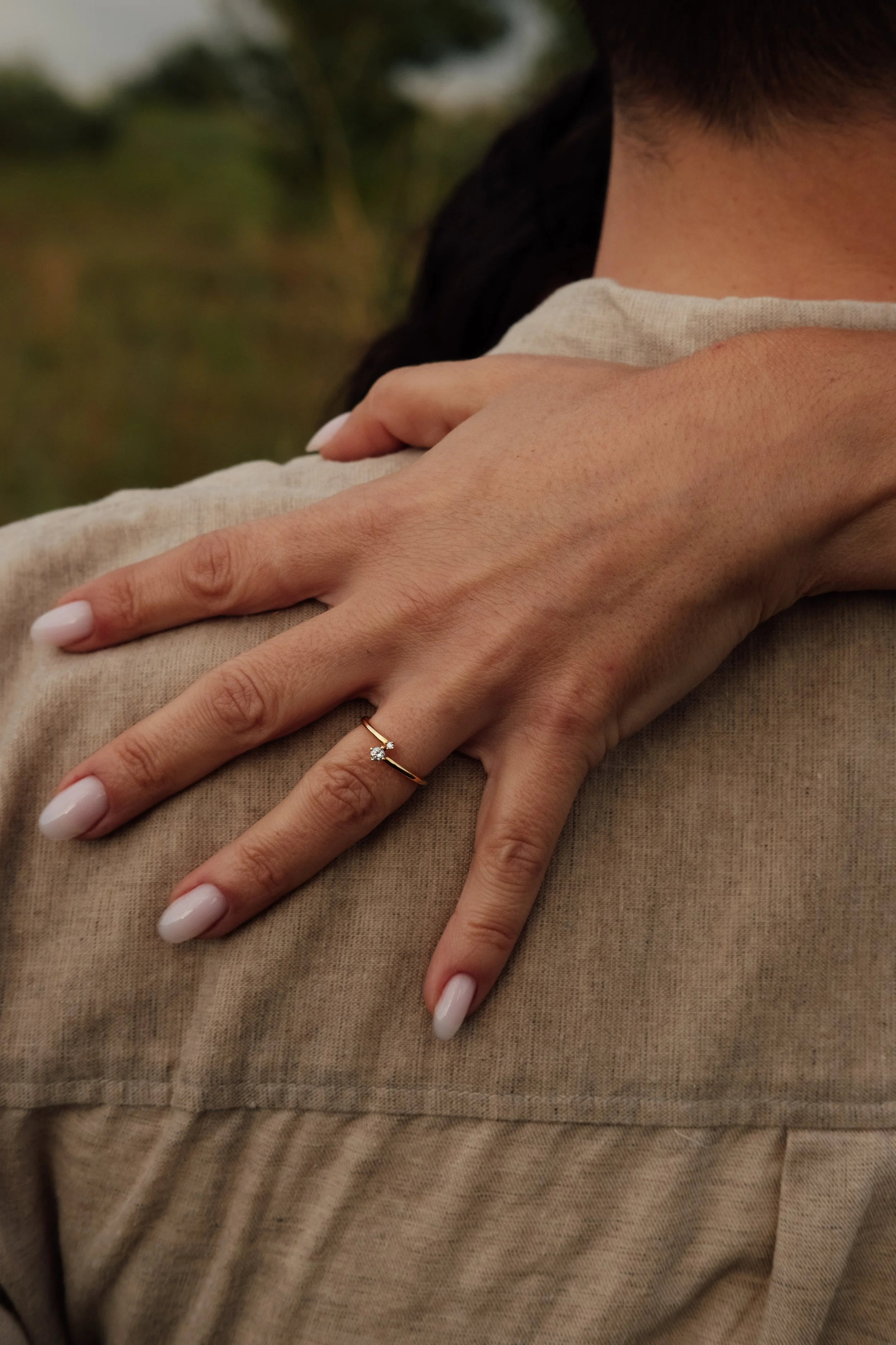 A close-up of a person hugging another person, with their hand resting on the other's shoulder. The person wearing the ring has white, manicured nails and light skin. They are dressed in a beige or light brown shirt. The background is outdoors with b