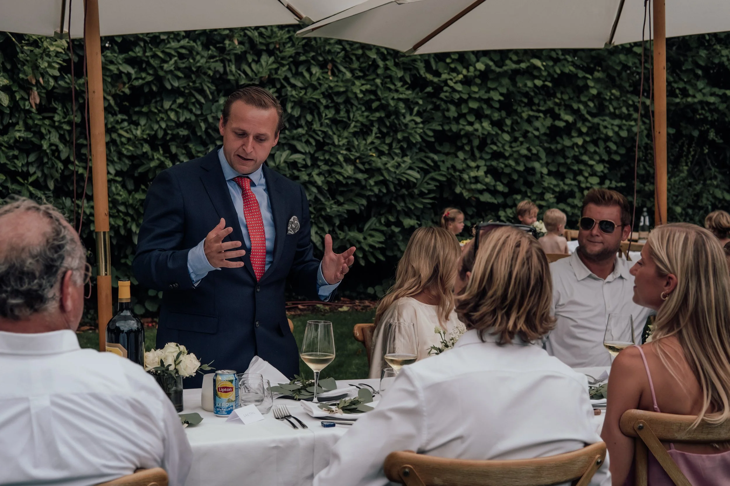 Man in a blue suit and red tie speaking at a dinner party outdoors under umbrella, with guests seated at tables with white tablecloths and floral centerpieces.