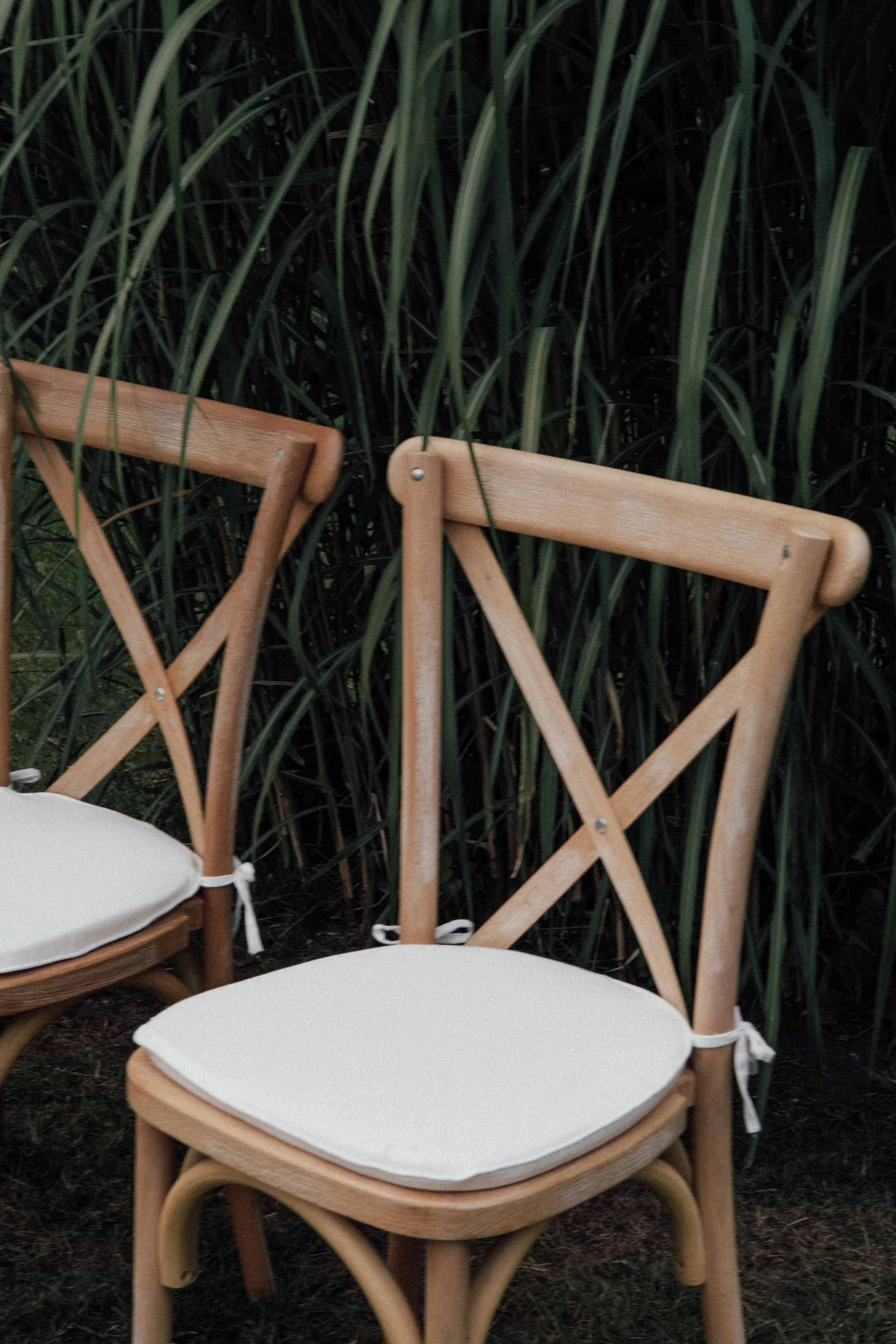 Multiple wooden and rattan chairs with white cushions arranged outdoors in front of tall green grass.