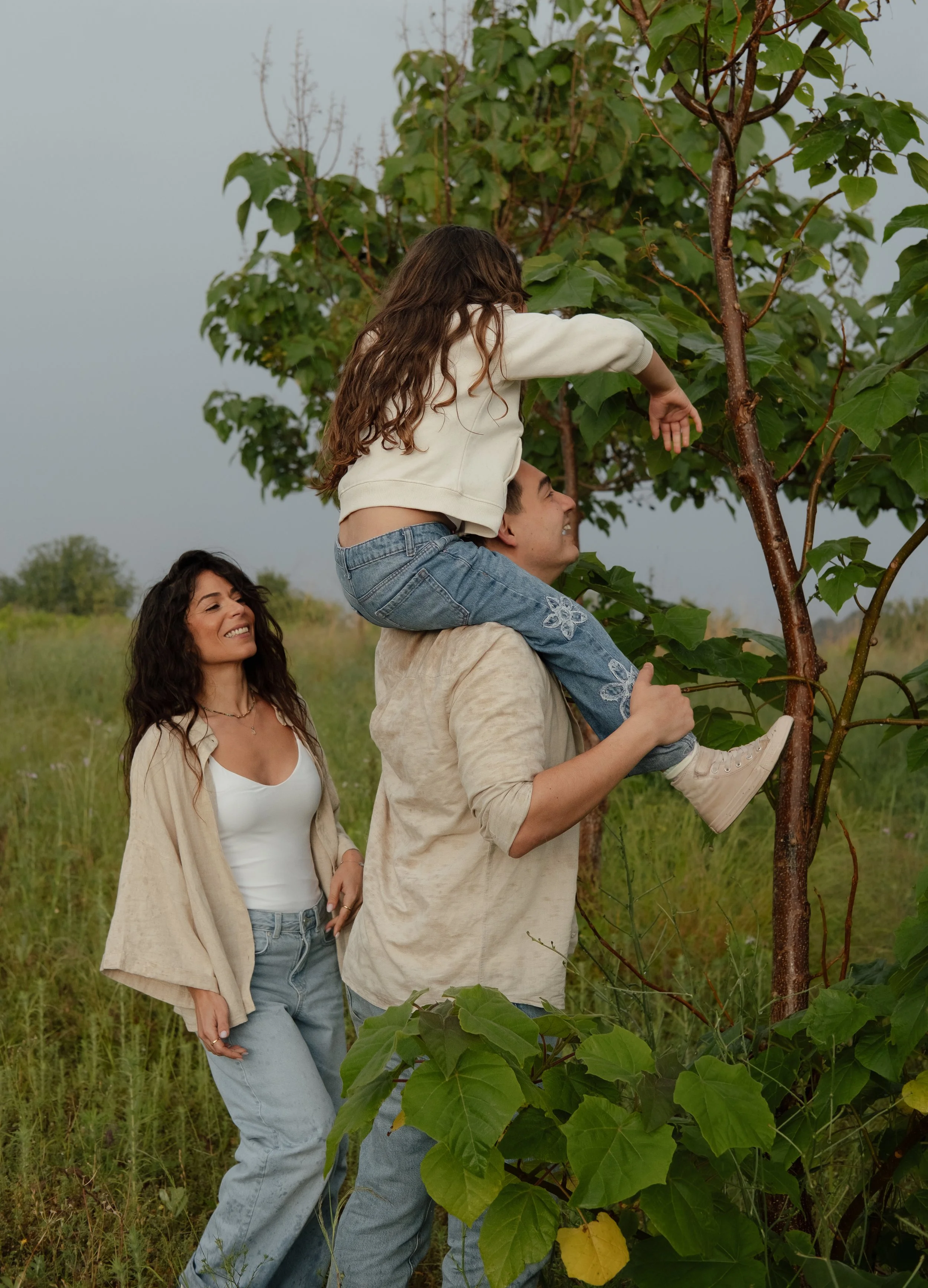 A man is holding a young girl on his shoulders as they pick leaves from a tree in an open field. A woman is standing nearby, smiling and observing their activity.