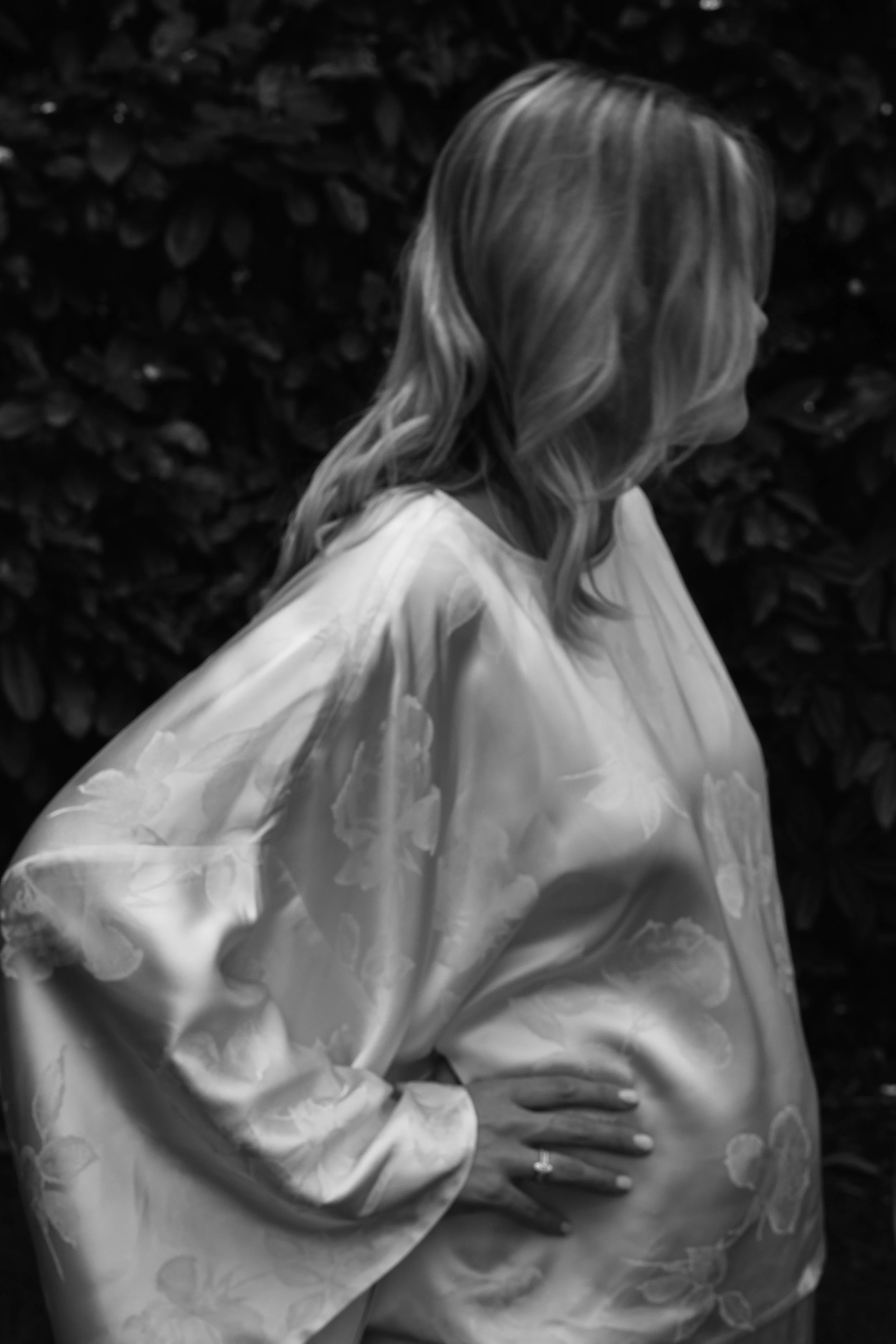 A person dressed in a shiny satin gown with floral patterns holding their hands together, standing against a dark, leafy background.