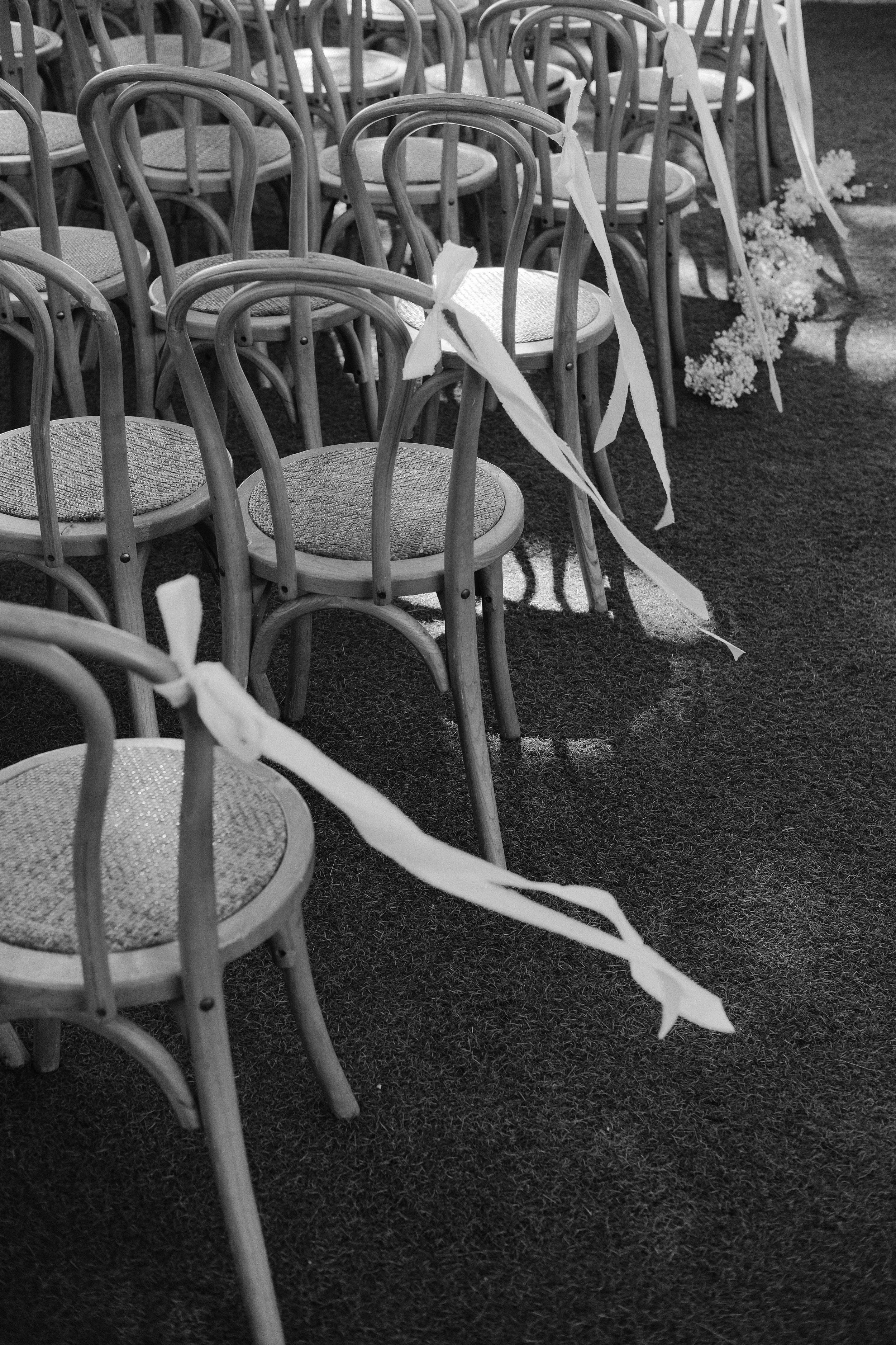 Empty wooden chairs with woven seats decorated with paper ribbons and flowers, arranged outdoors on grass, possibly for an event or gathering.