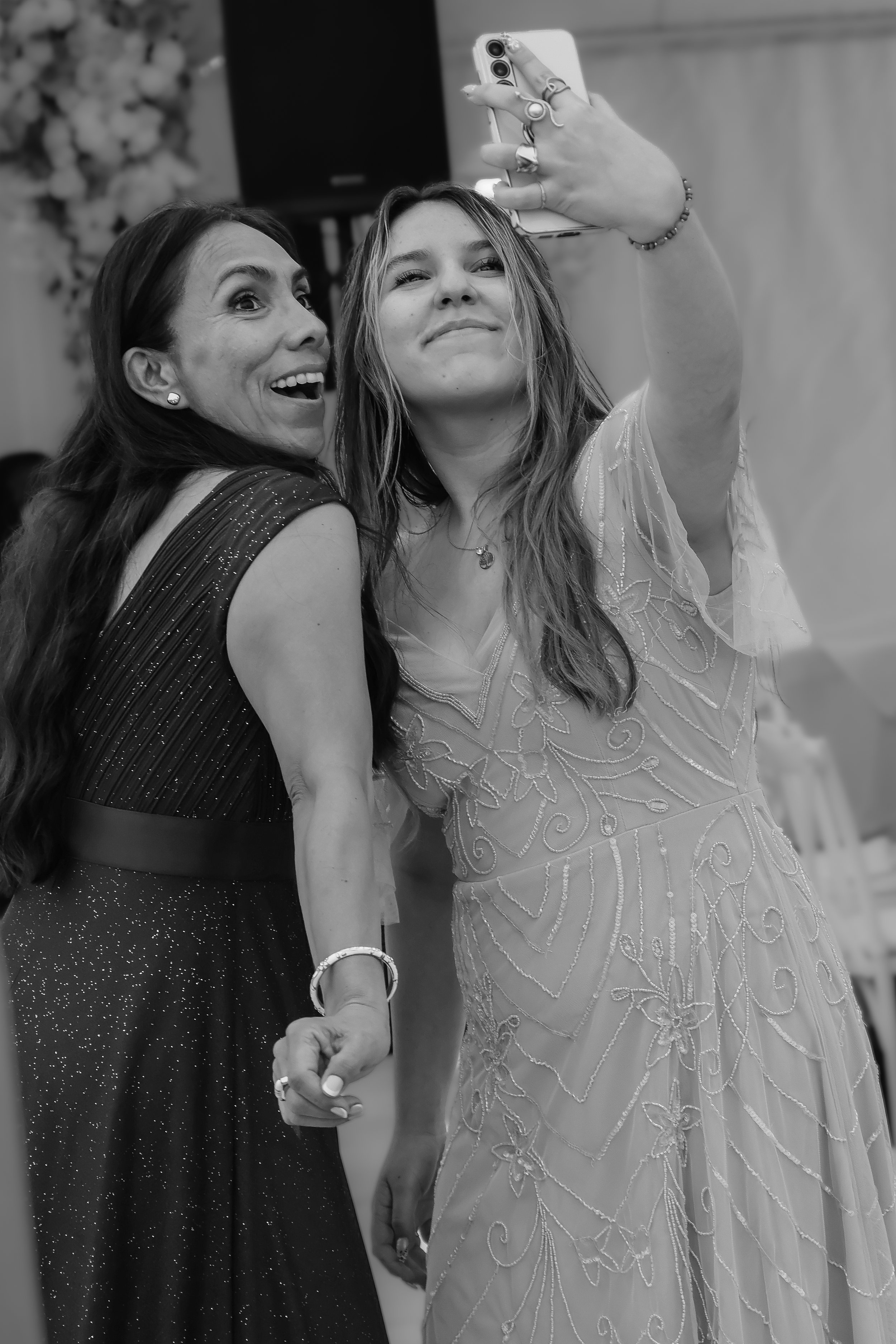 Two women taking a selfie together at a celebratory event, holding hands and smiling. One woman is wearing a sparkly dress, and the other is in a detailed embroidered gown.