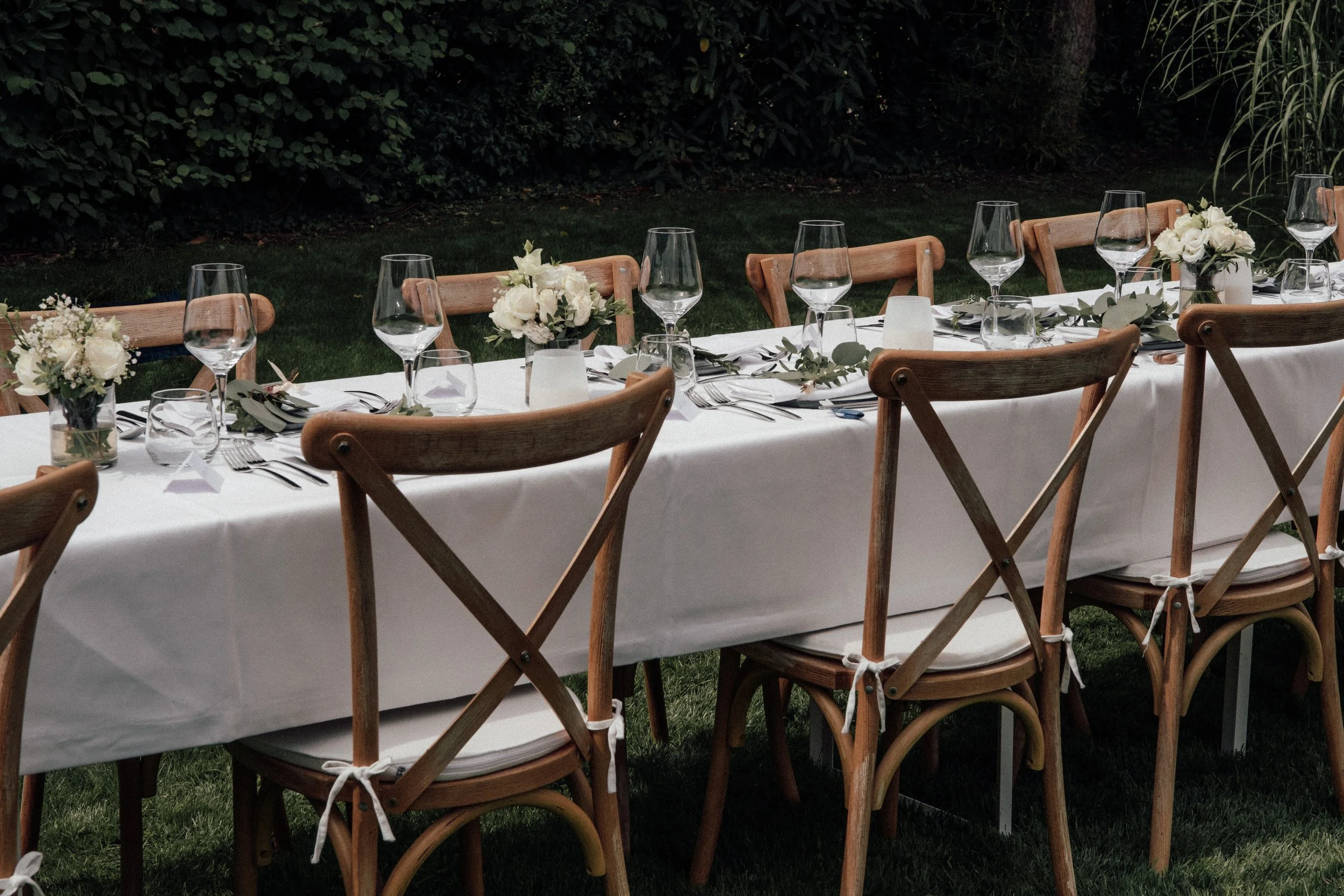 A long outdoor dining table set for a formal event with white tablecloth, floral centerpieces, glassware, and utensils, surrounded by wooden chairs, in a garden setting at dusk.