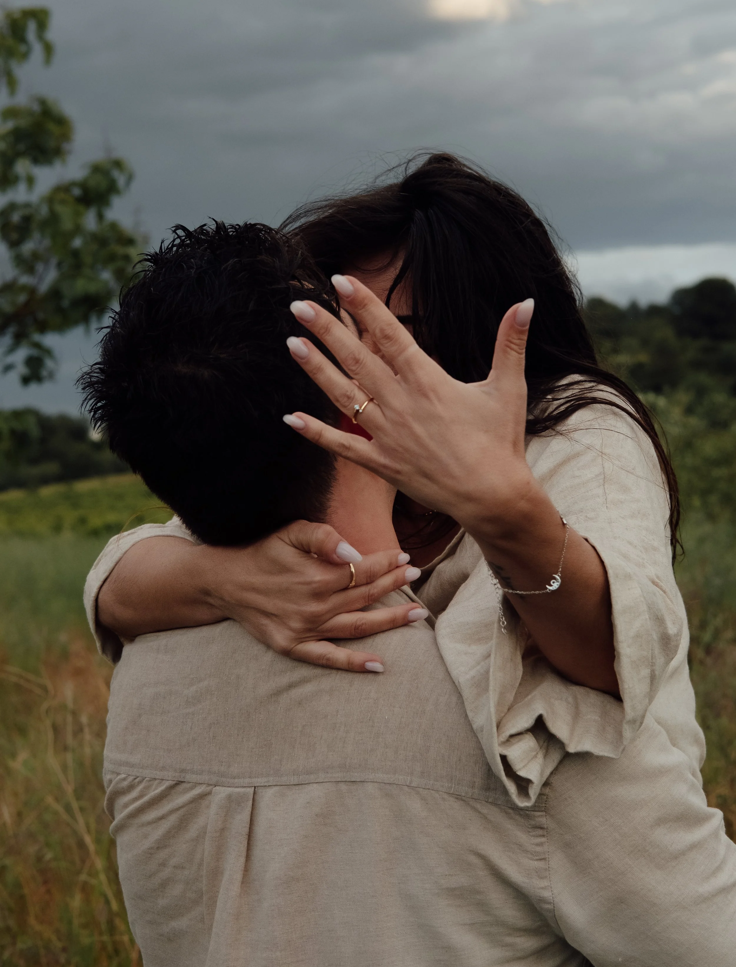 A couple embracing outdoors, the woman holding the man's head while they kiss, with trees and cloudy sky in the background.