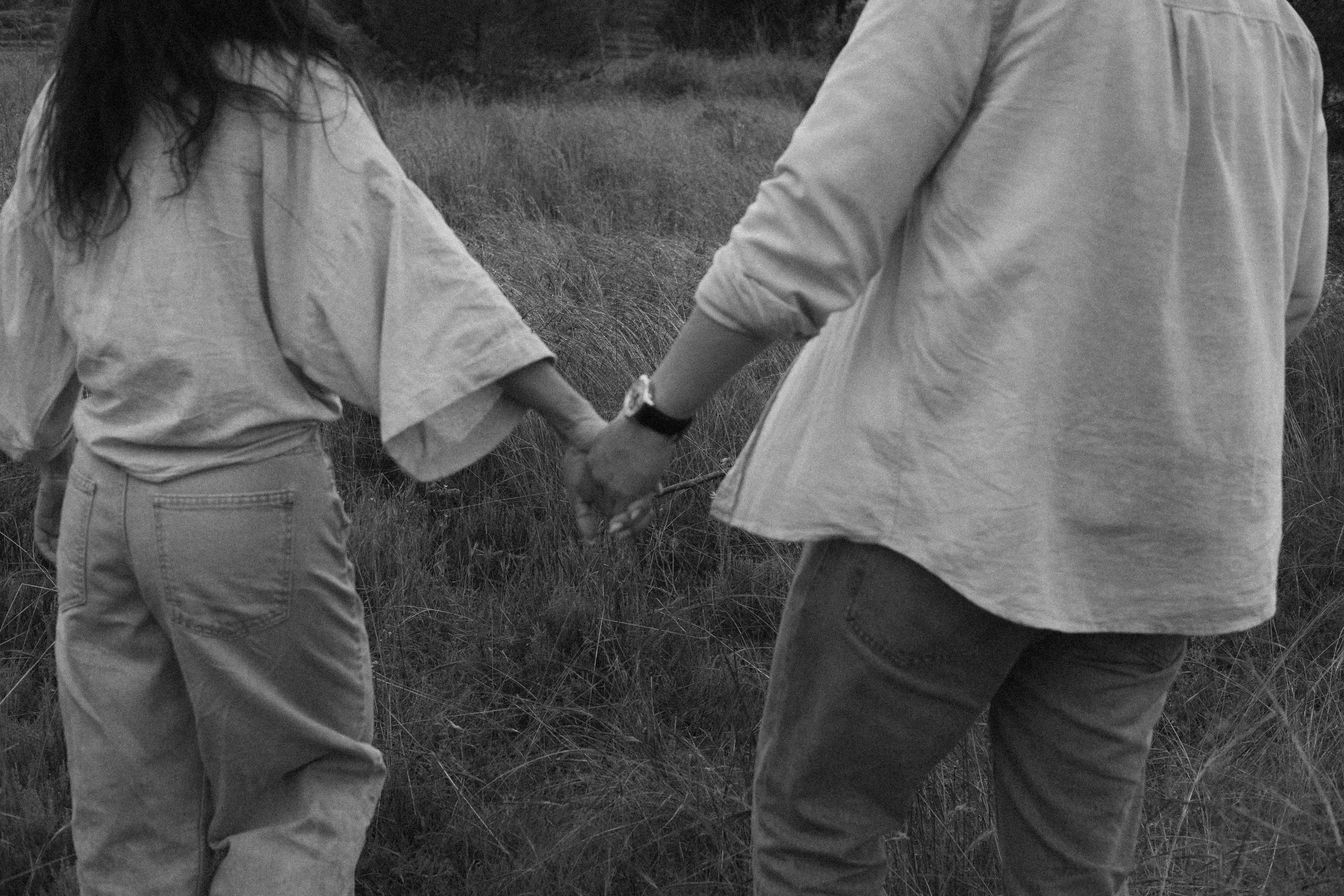 Two people holding hands in a grassy outdoor area, seen from the waist up, wearing casual clothing.