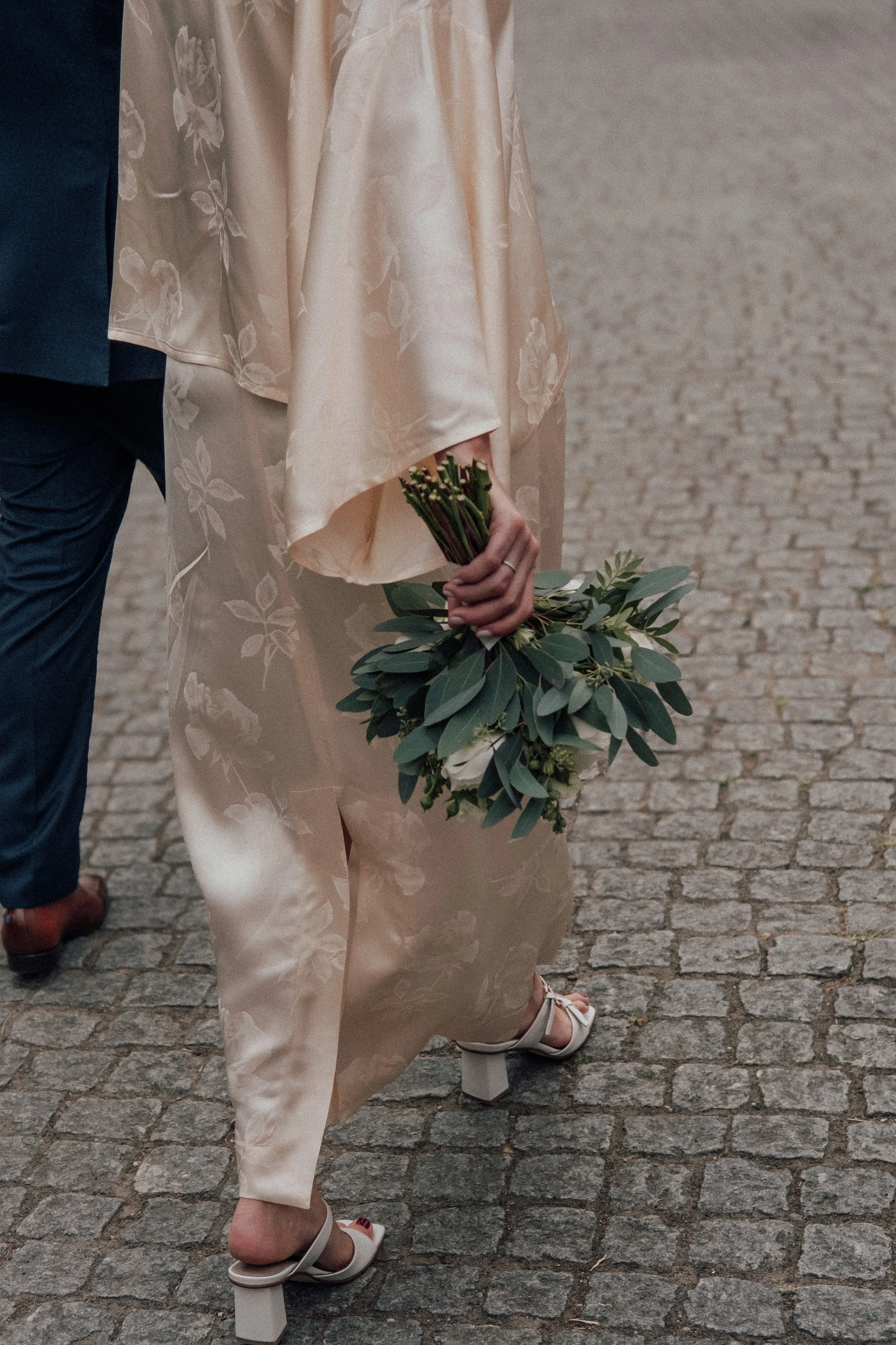 Person wearing a beige floral dress and white high-heeled sandals holding a bouquet of greenery, walking on cobblestone street, next to a person in dark blue pants and shoes.