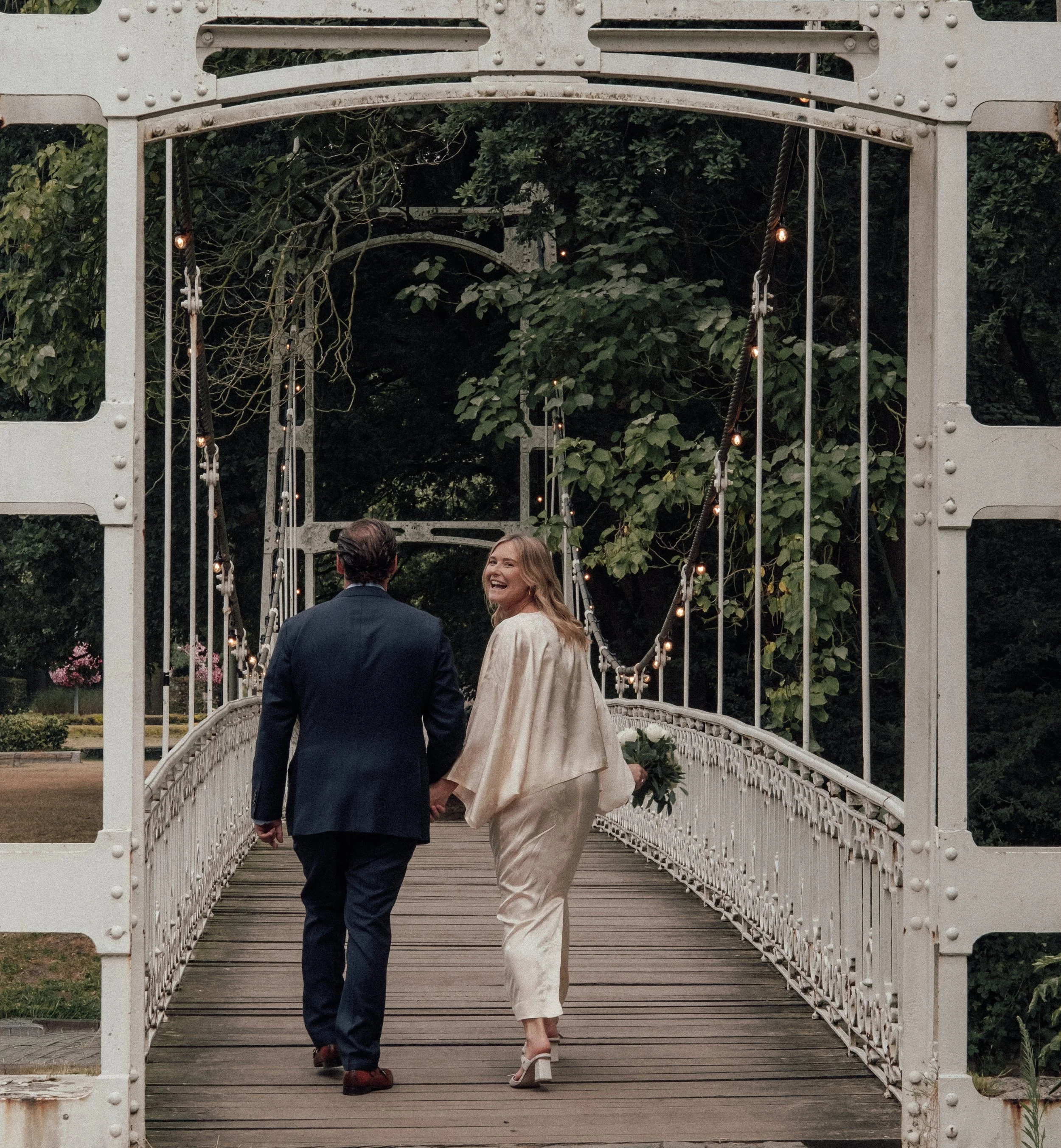 A man and a woman walk hand in hand through a white metal bridge, with lush green trees in the background. The woman is smiling and wearing a cream-colored dress with high heels, while the man is dressed in a navy suit.