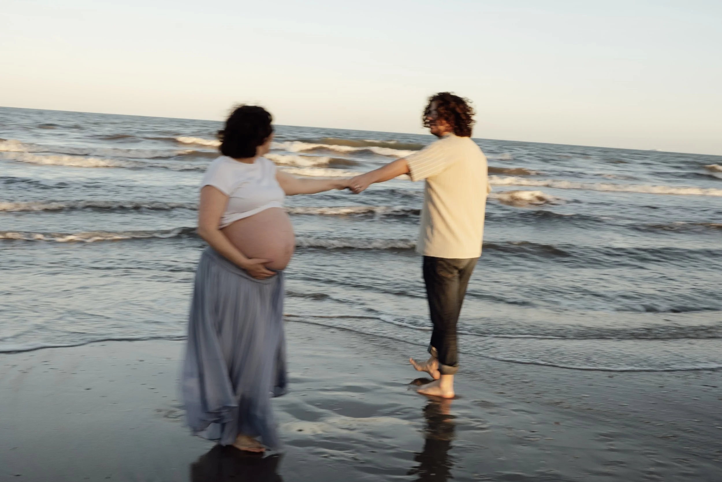 Pregnant couple holding hands while walking barefoot along the beach at sunset