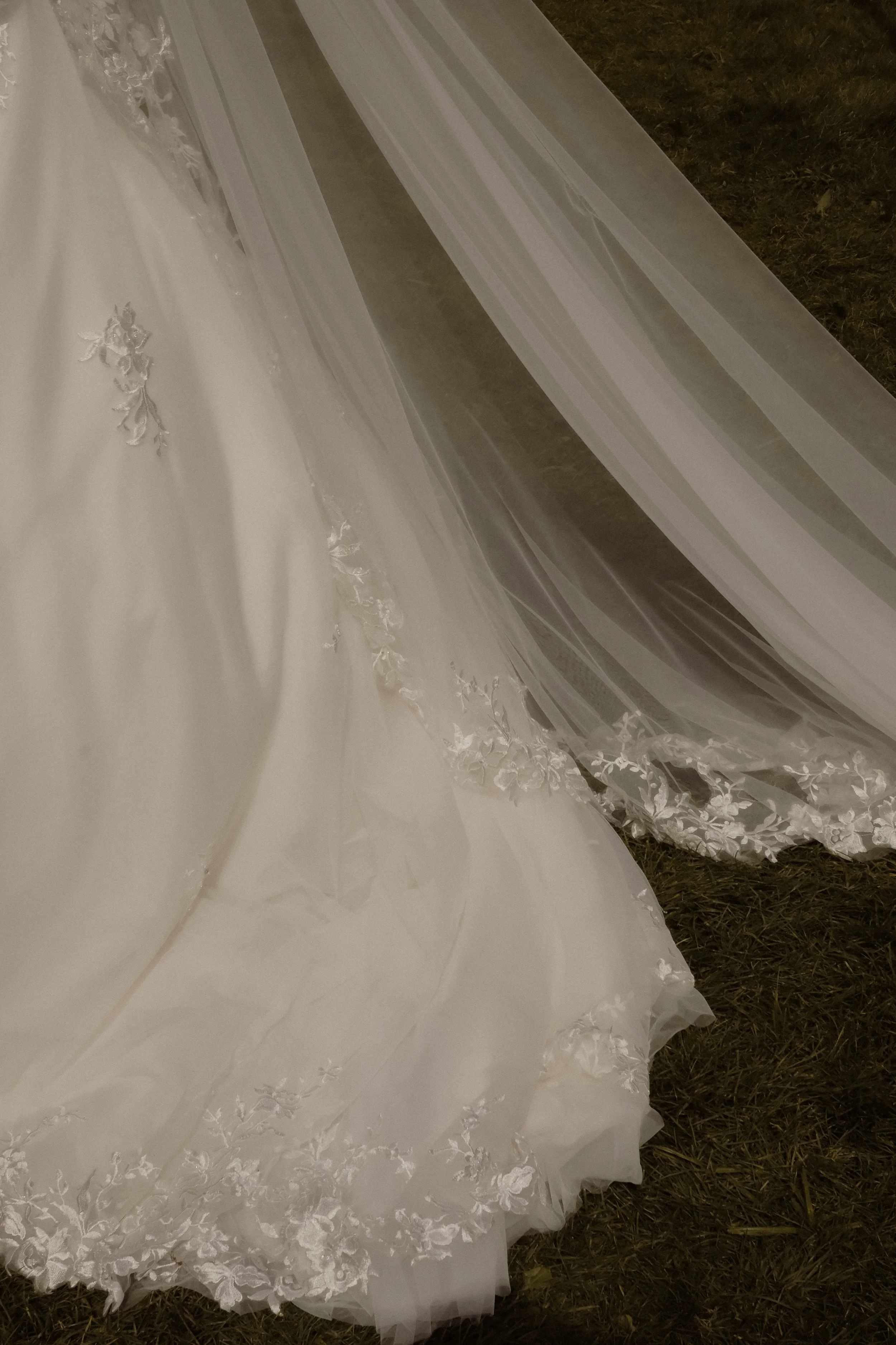 Close-up view of white lace and tulle fabric, likely part of a wedding dress, laid on grass.