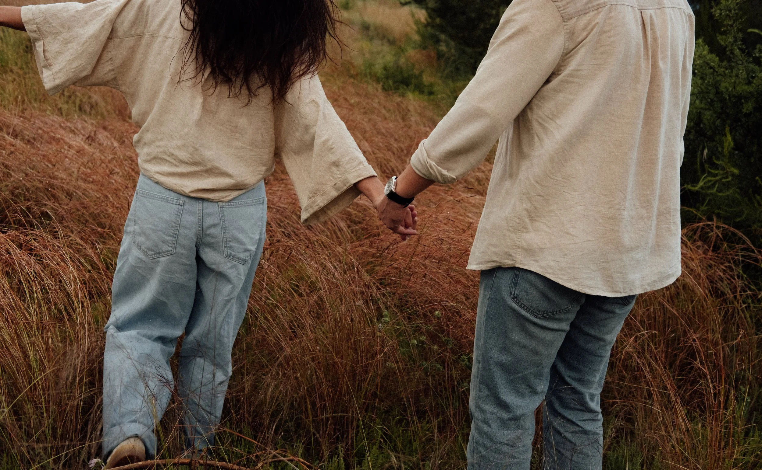 Two people holding hands while walking through tall grass in a natural outdoor setting.